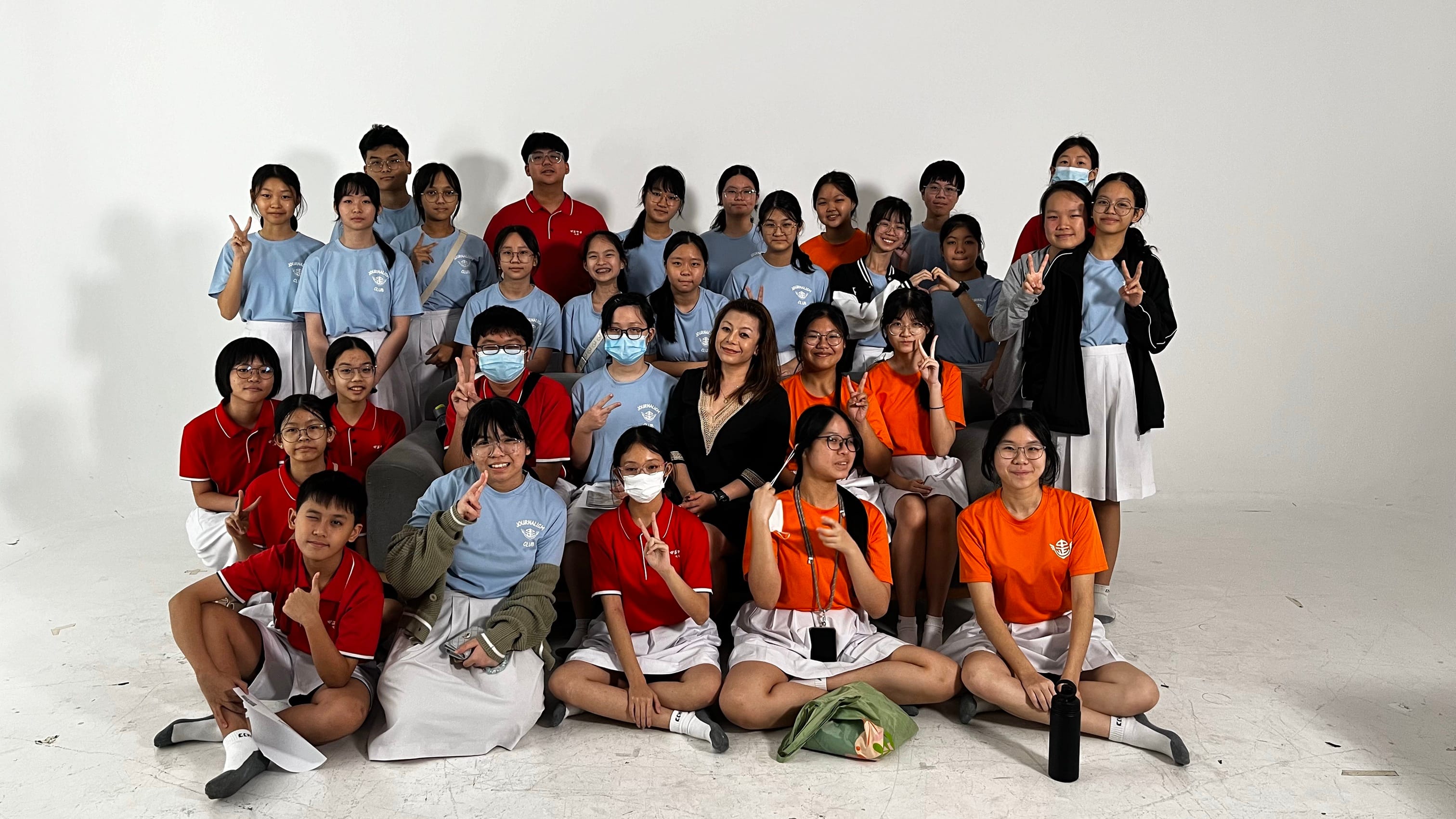 A large group of students in blue, red, and orange uniforms pose with a teacher against a white background.