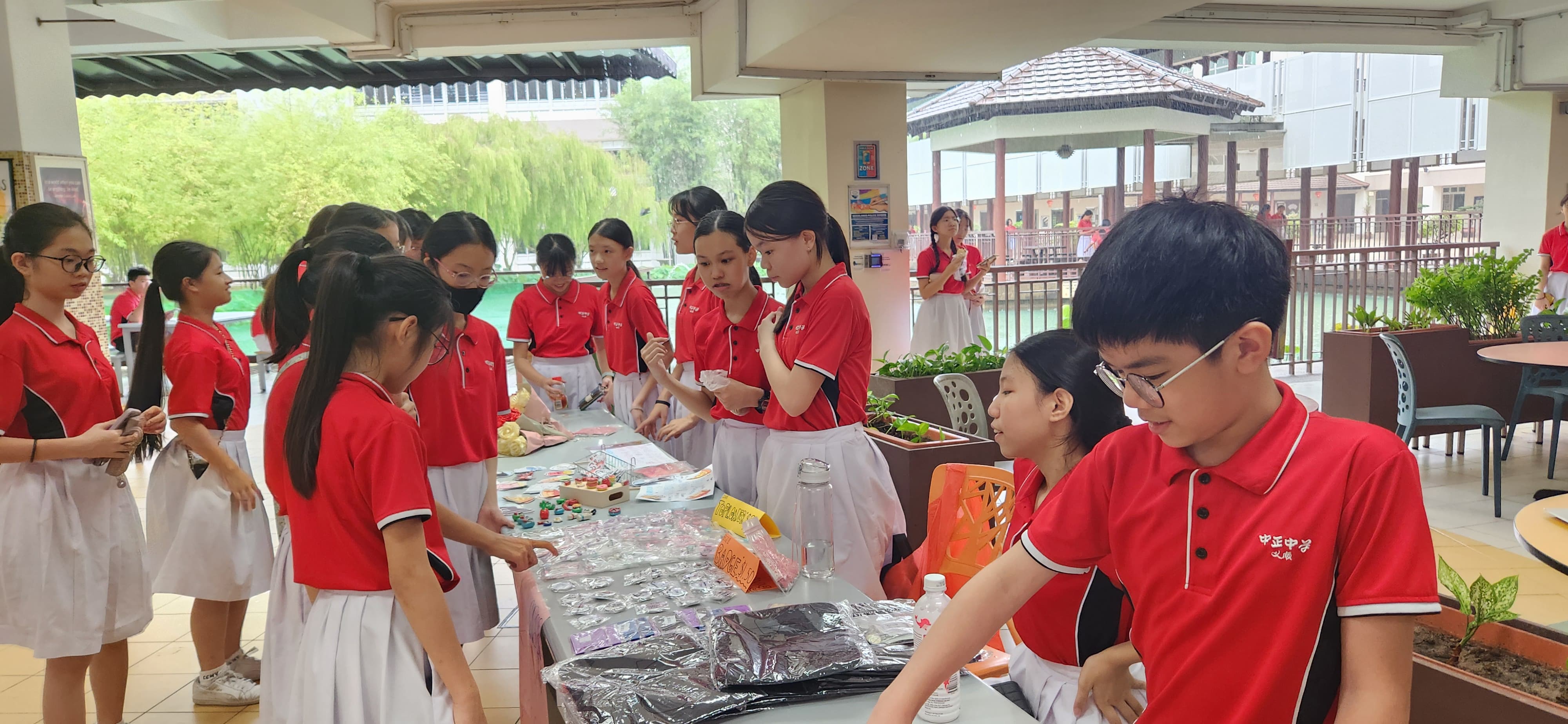 Students in red and white uniforms gather around a table displaying various small items at an outdoor school event.