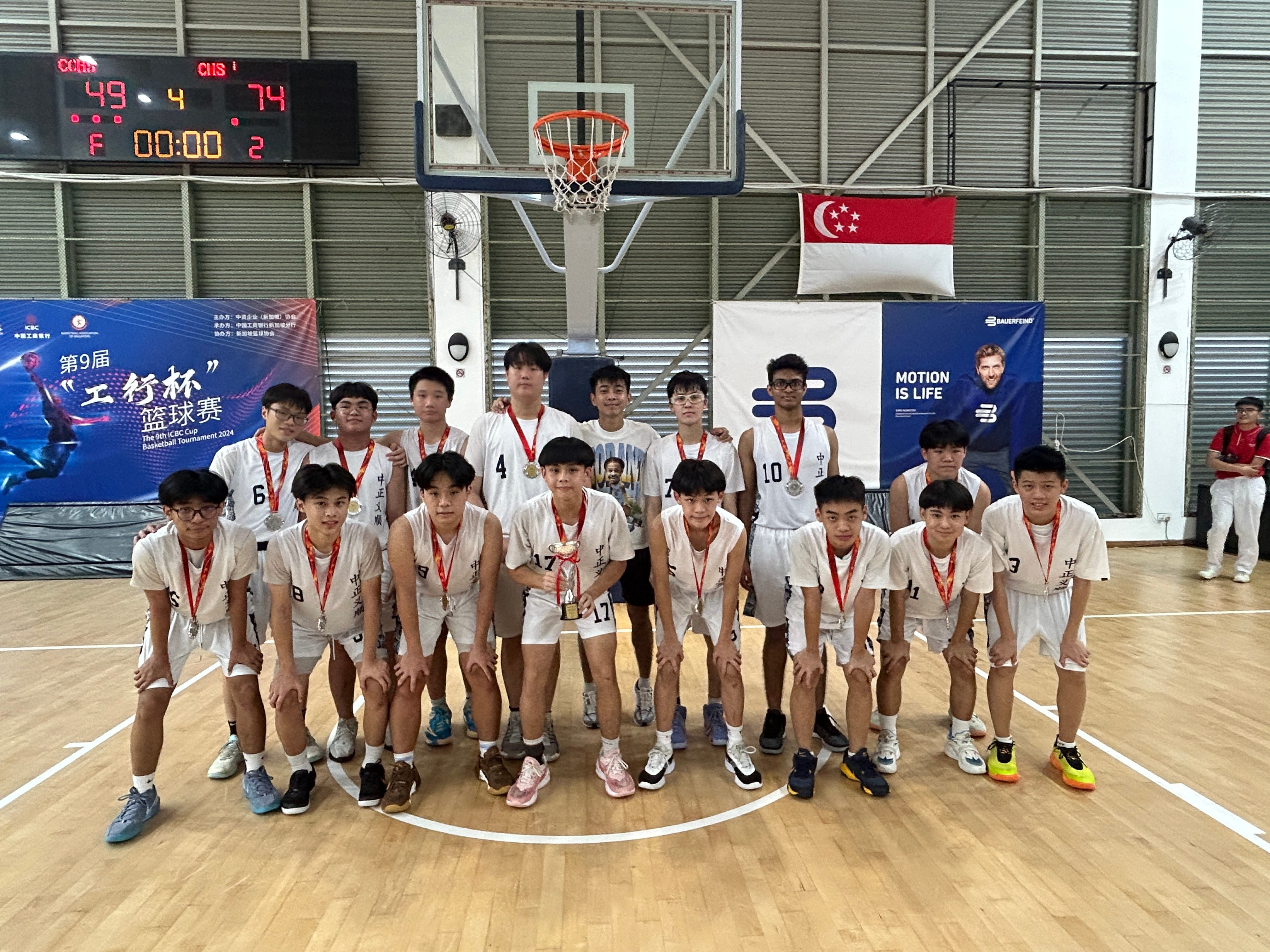 A youth basketball team in white uniforms poses with medals and a trophy on an indoor court after winning a tournament.