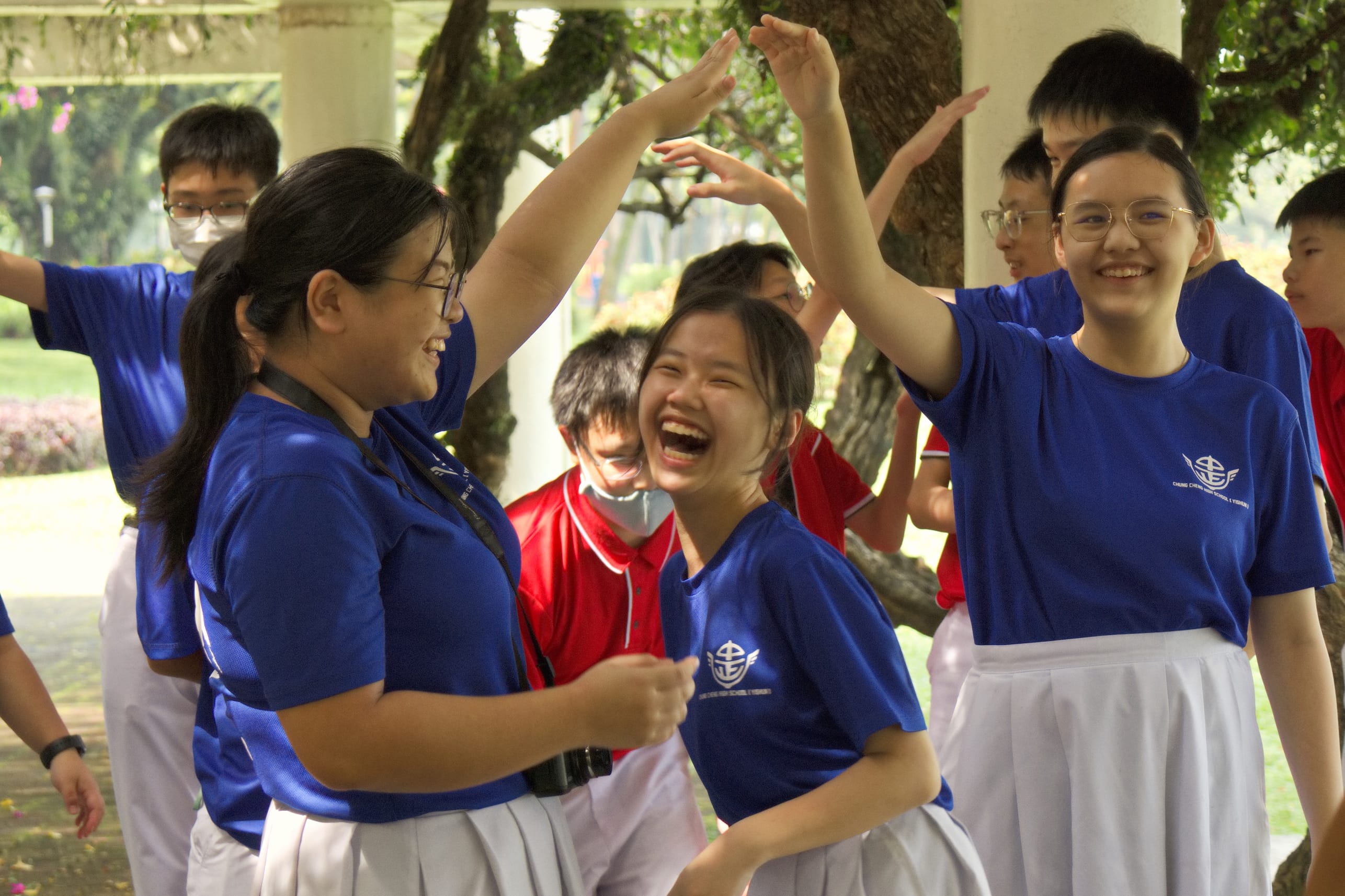 Students in blue and white uniforms laugh and raise hands together outdoors near trees and a covered walkway.