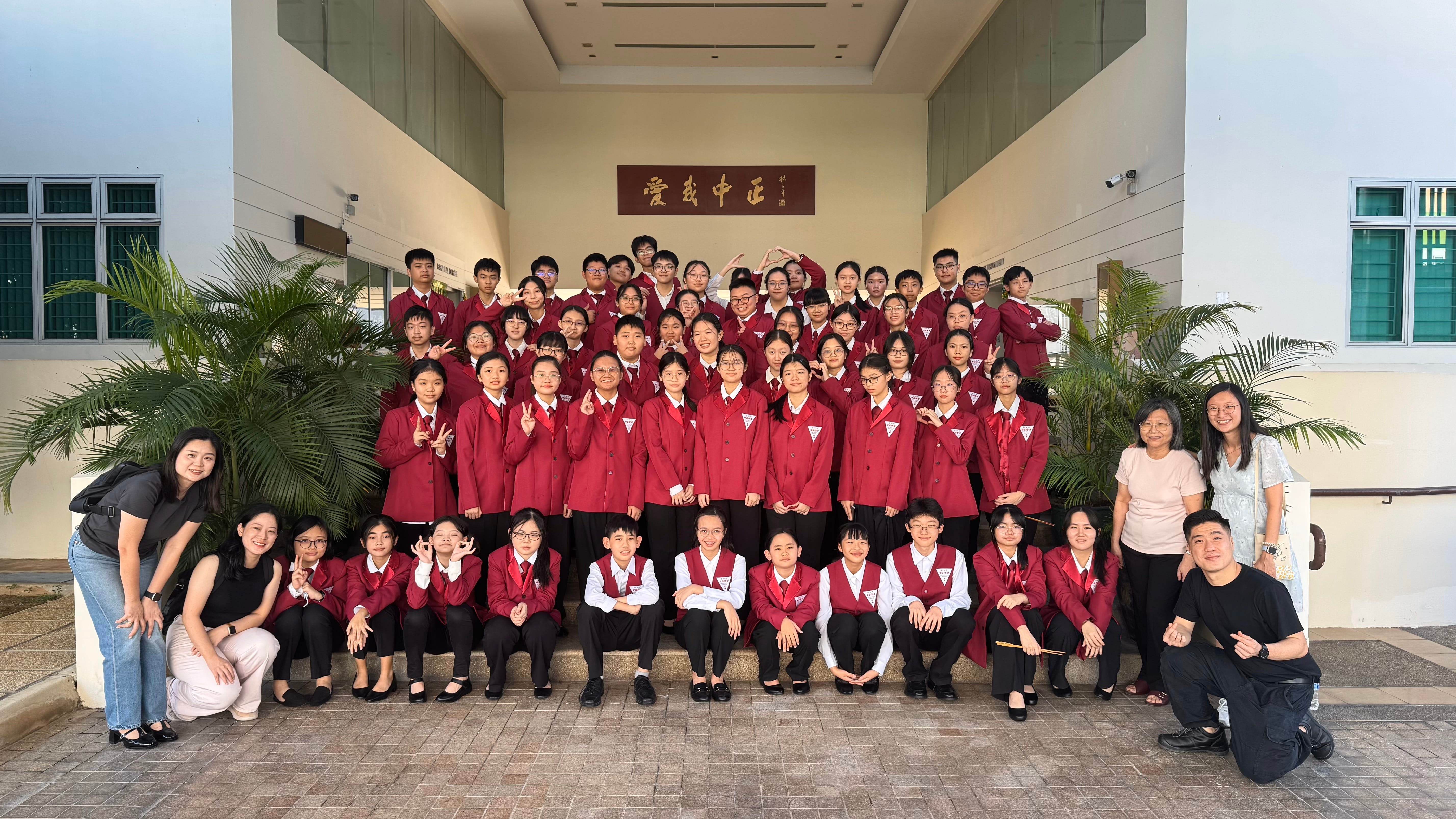 Large group in matching red blazers pose on steps between plants in a school courtyard, with teachers at sides.