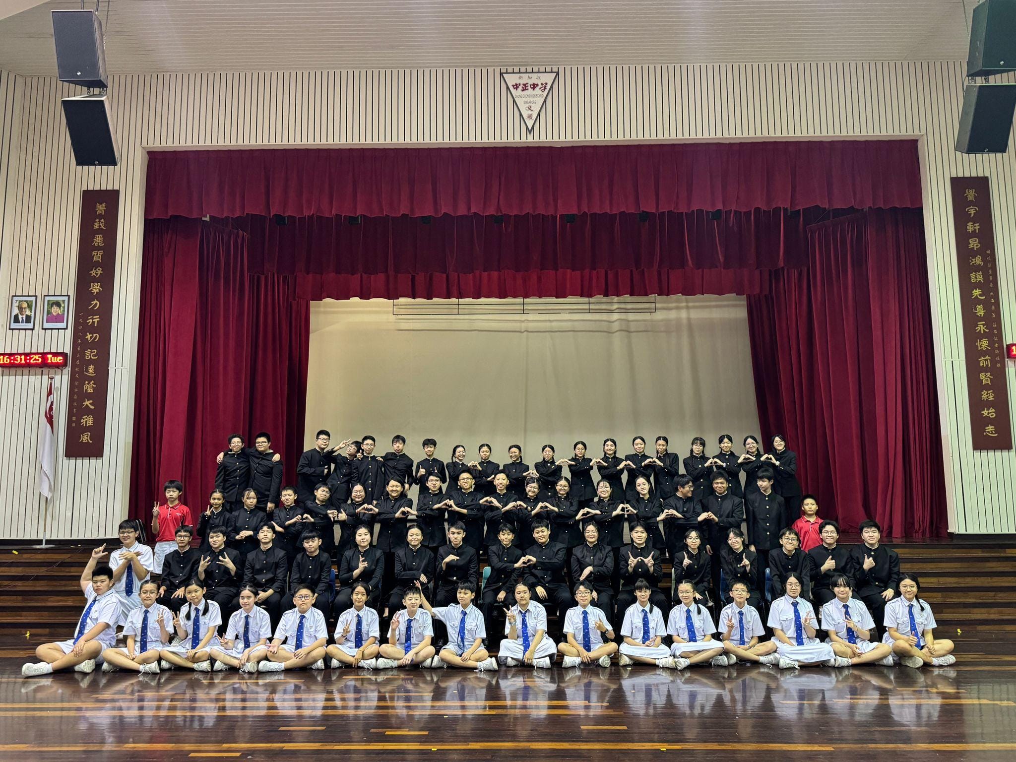 Large group of students in black and white uniforms posing together in a school hall with red curtained stage.