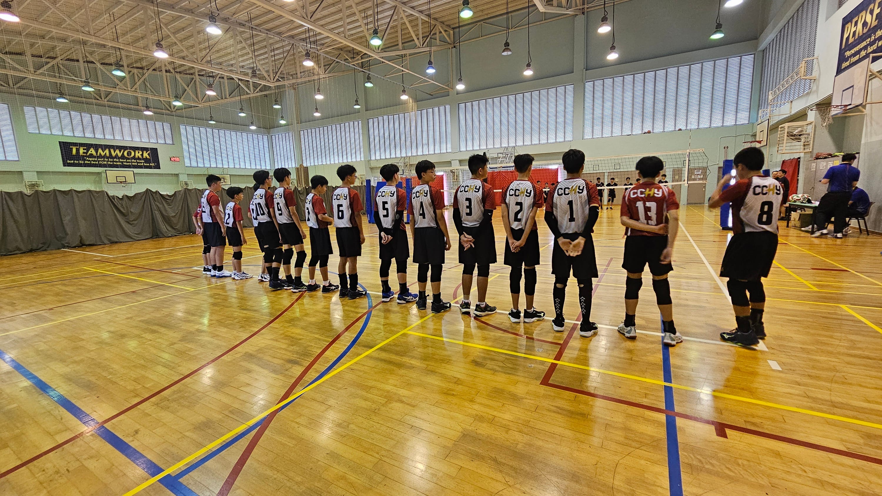 A row of young male volleyball players in numbered CCHY jerseys stand in an indoor gymnasium with a Teamwork banner.