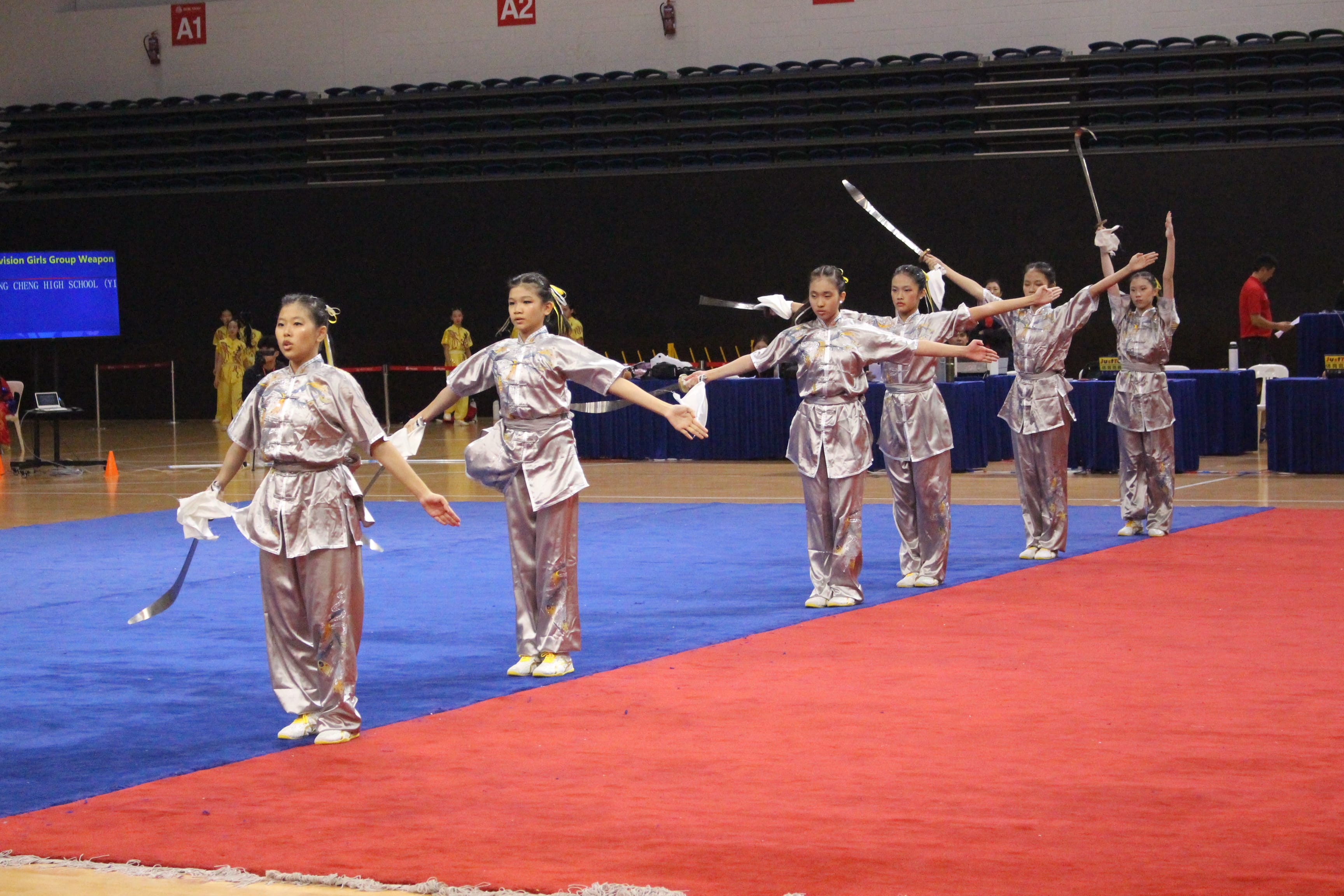 Six girls in shiny silver outfits perform a synchronized routine with swords on a blue and red mat.
