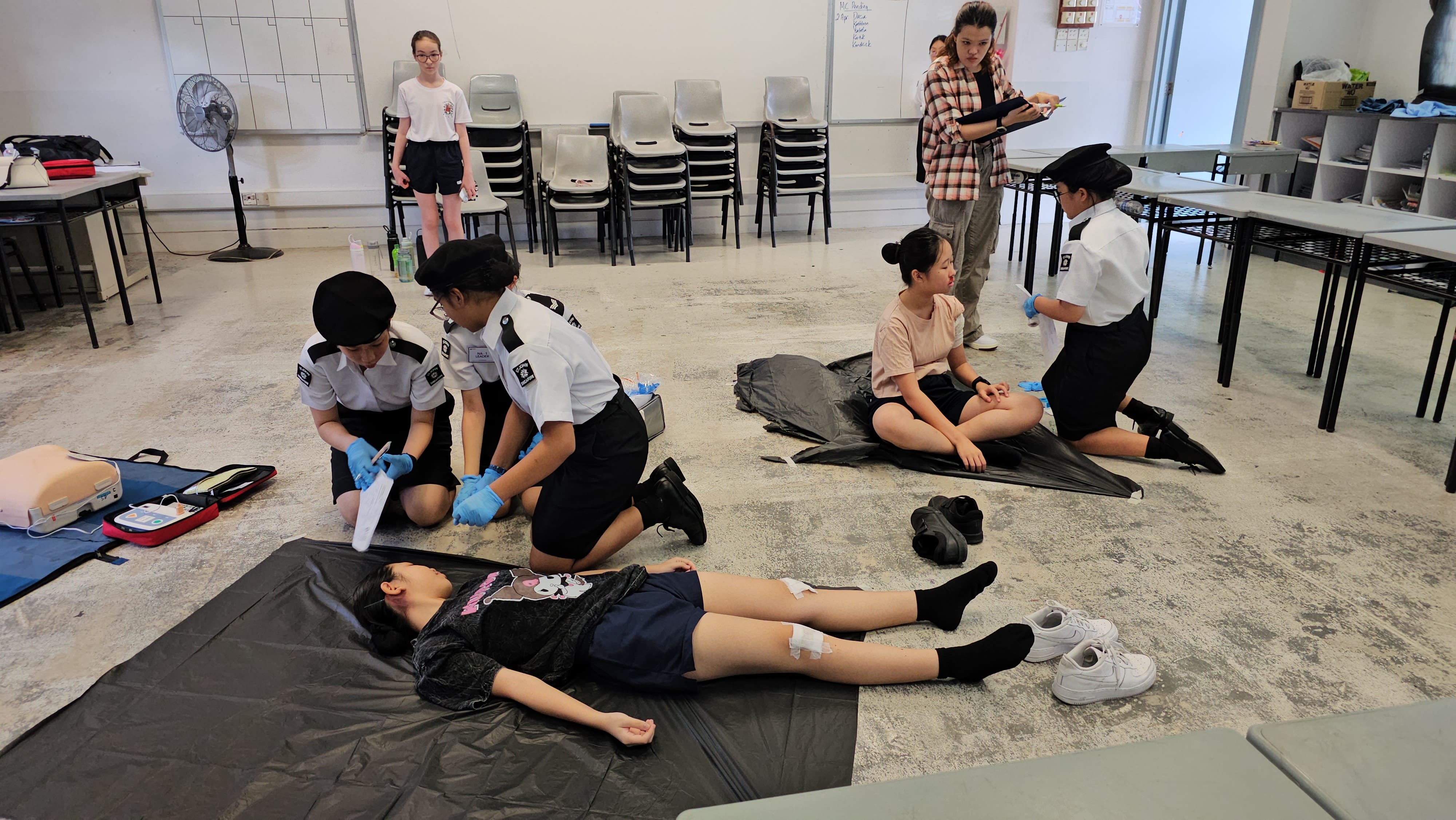 Students in uniform practice first aid on peers lying on mats in a classroom, with an instructor observing nearby.