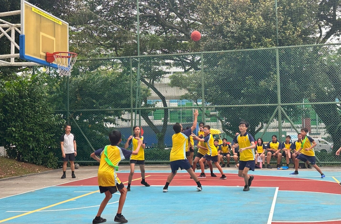 Students in yellow jerseys play basketball on an outdoor court, with one player shooting toward the hoop.