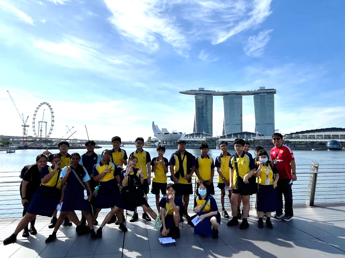 Students in yellow and blue uniforms pose by the waterfront with Marina Bay Sands and the Flyer in the background.