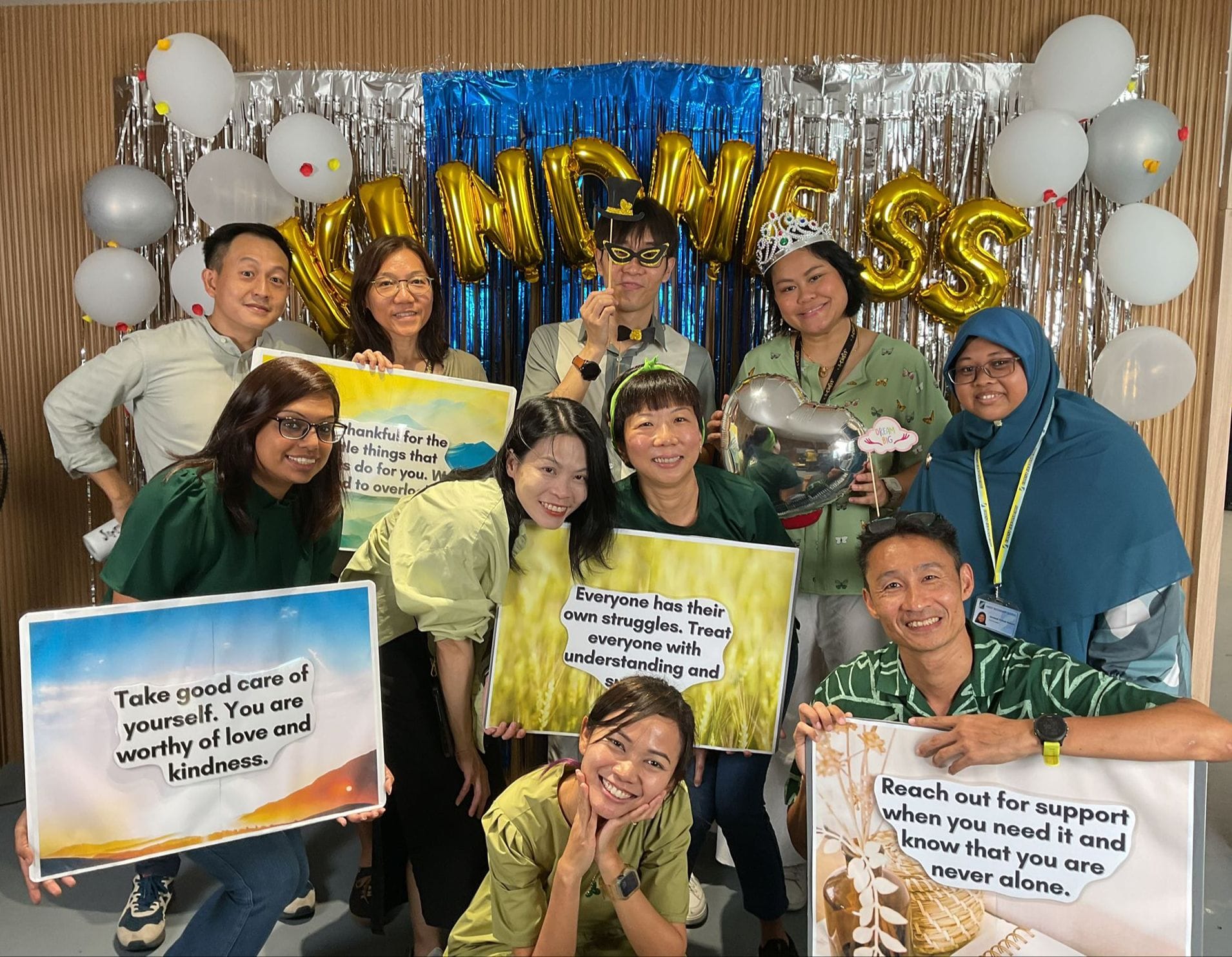 Group of people posing with kindness-themed signs in front of a gold balloon backdrop spelling KINDNESS.