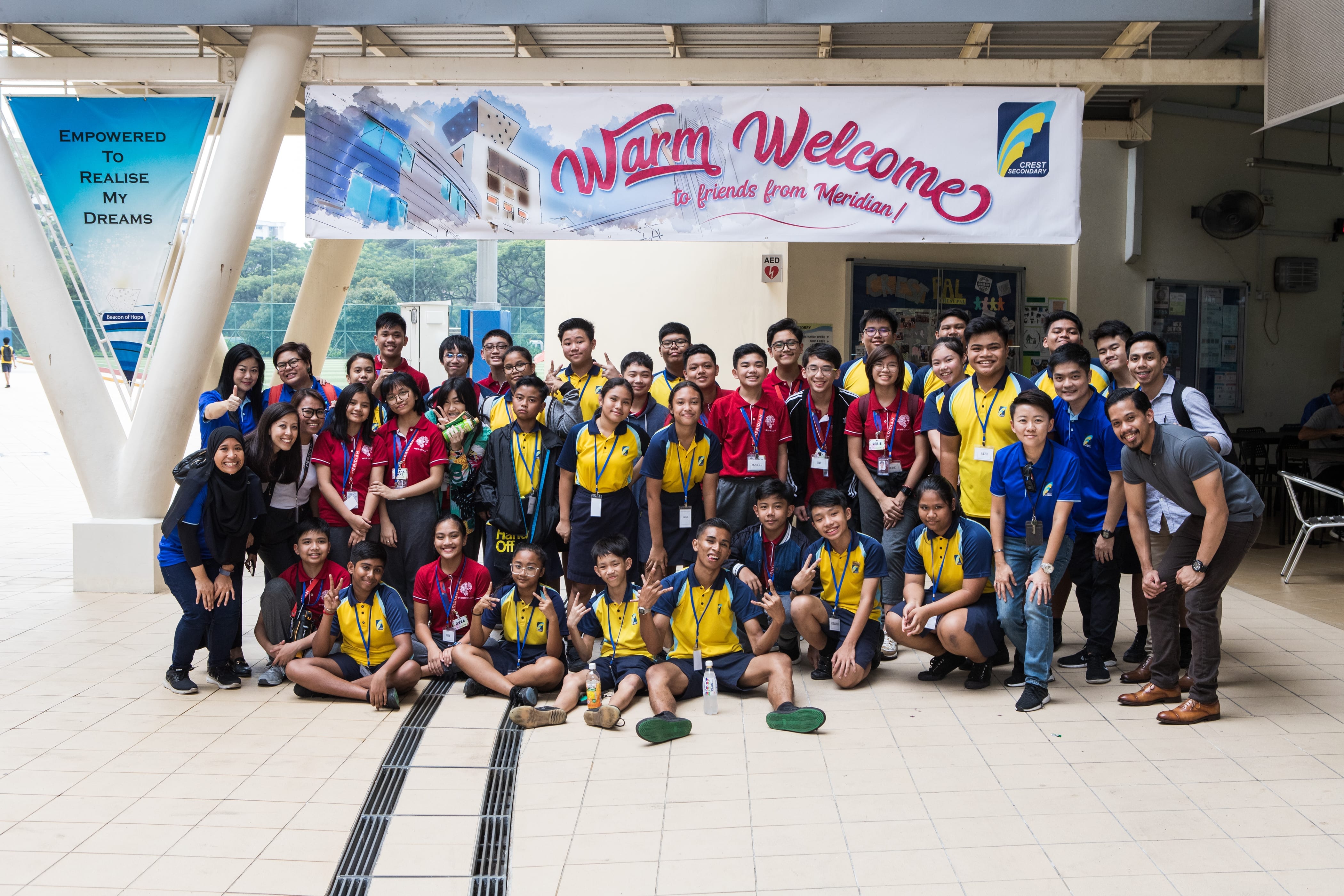 A large group of students and teachers pose under a banner reading Warm Welcome to friends from Meridian at Crest Secondary.