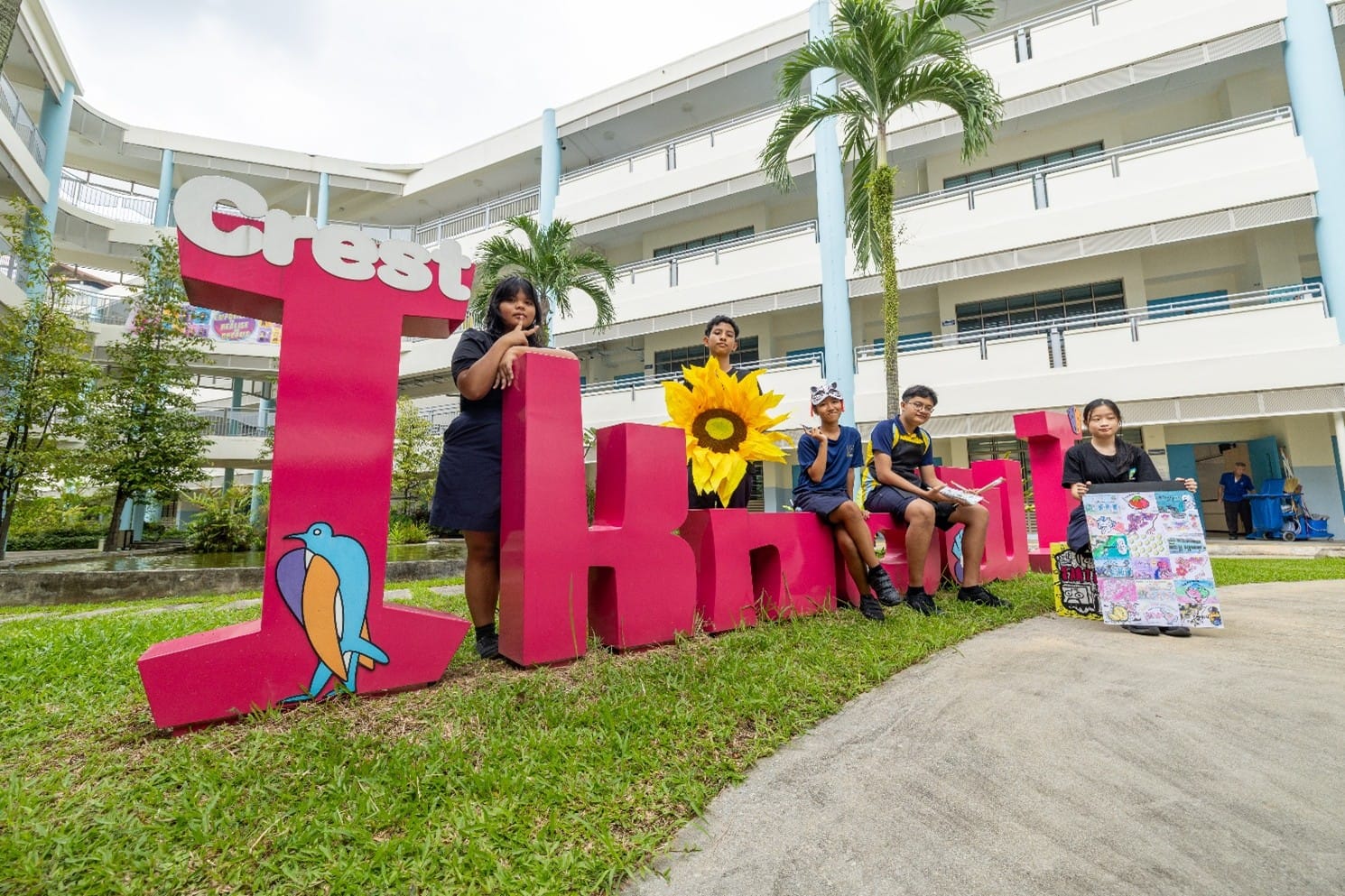 Four students pose with large pink "Crest" letters on grass in a school courtyard with palm trees.