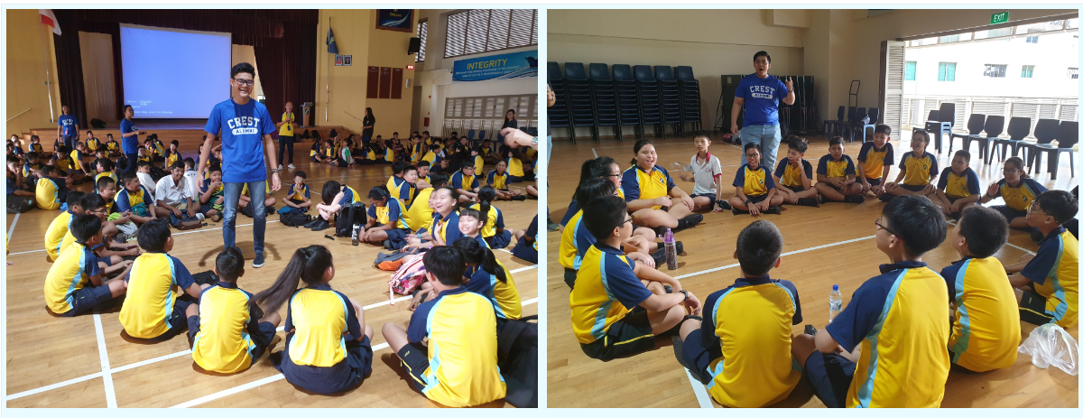 A man in a blue Crest shirt stands and speaks to students in yellow uniforms sitting on a gym floor.