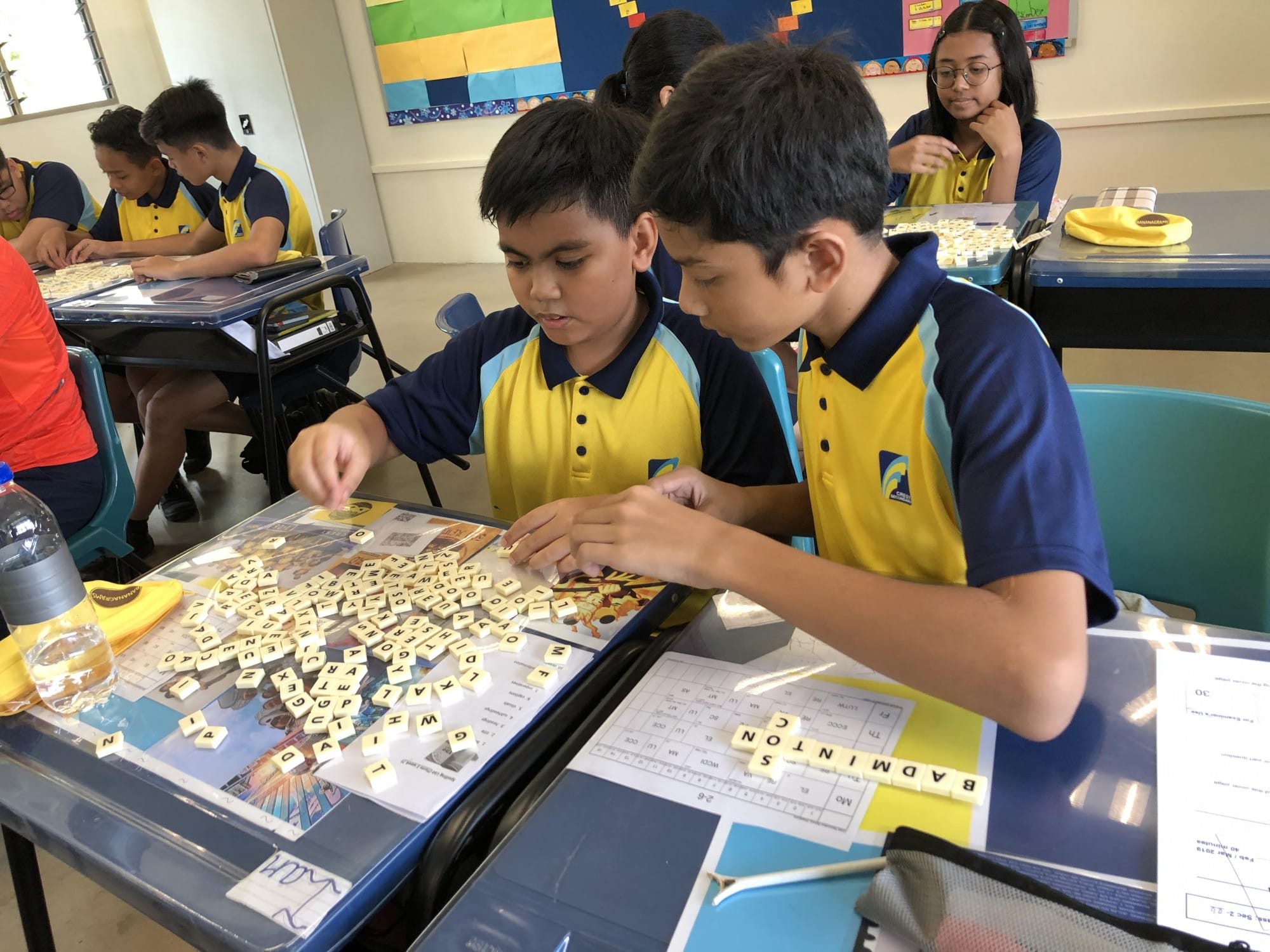 Two students in blue and yellow uniforms arrange letter tiles on a desk in a classroom.