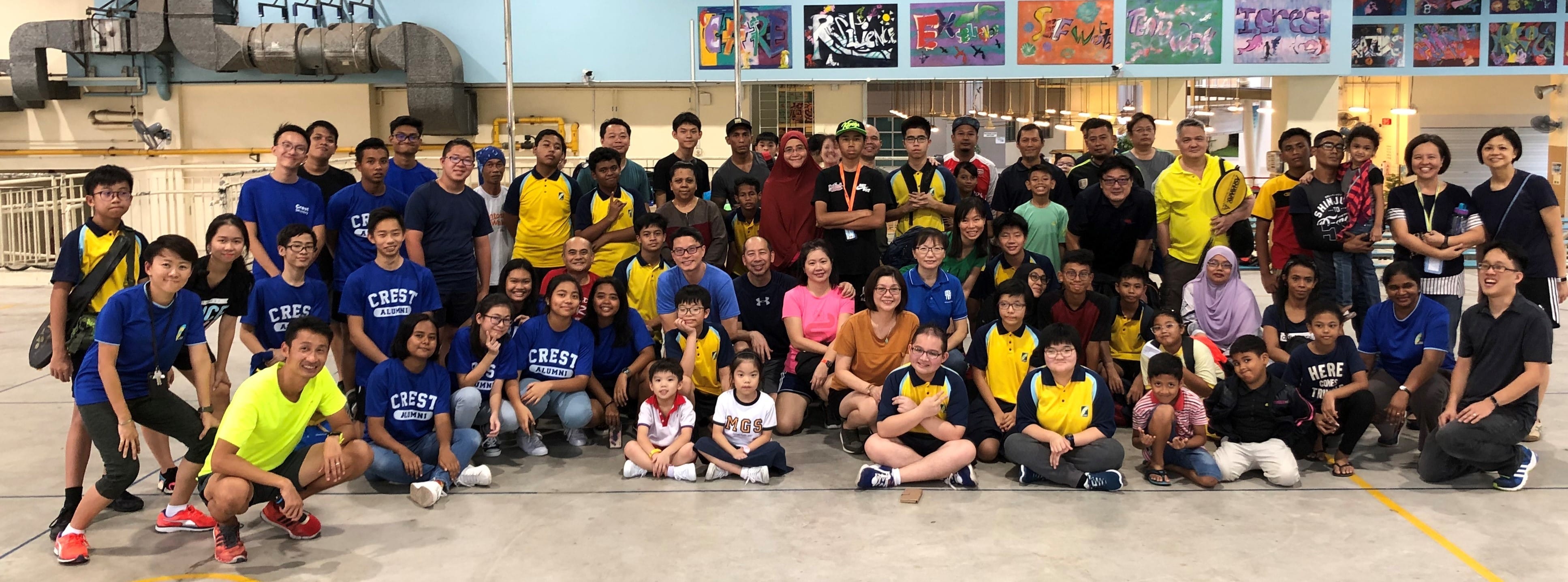 Large group of adults and children posing together indoors in a gymnasium with colorful artwork on the wall behind them.