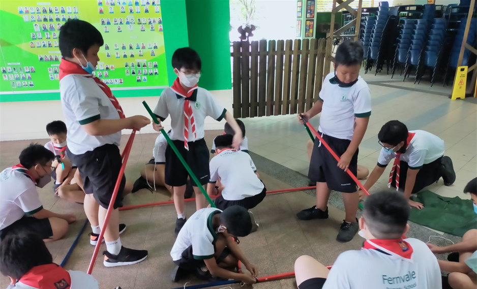 Students in uniforms and masks work together on the floor with colorful poles during a group activity indoors.