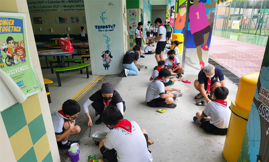 Students in scout uniforms with red neckerchiefs sit on a school corridor floor doing a hands-on activity with adults.