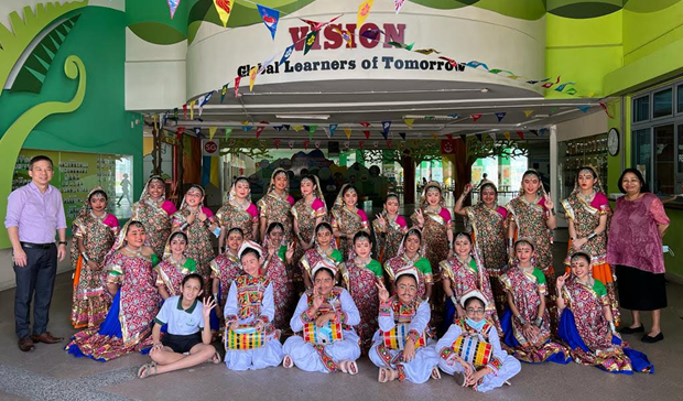 Group of children and adults in colorful traditional costumes posing under a "Vision: Global Learners of Tomorrow" sign.