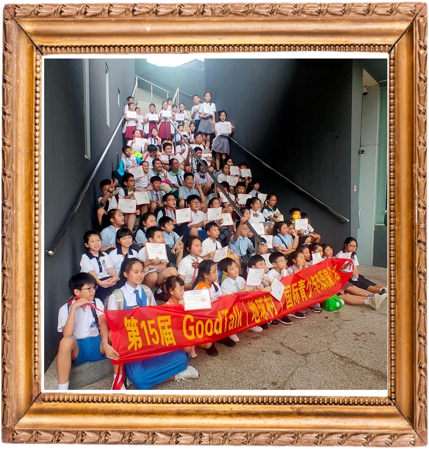 A large group of children on stairs holding certificates and a red Chinese banner reading 15th GoodTalk, in a gold frame.