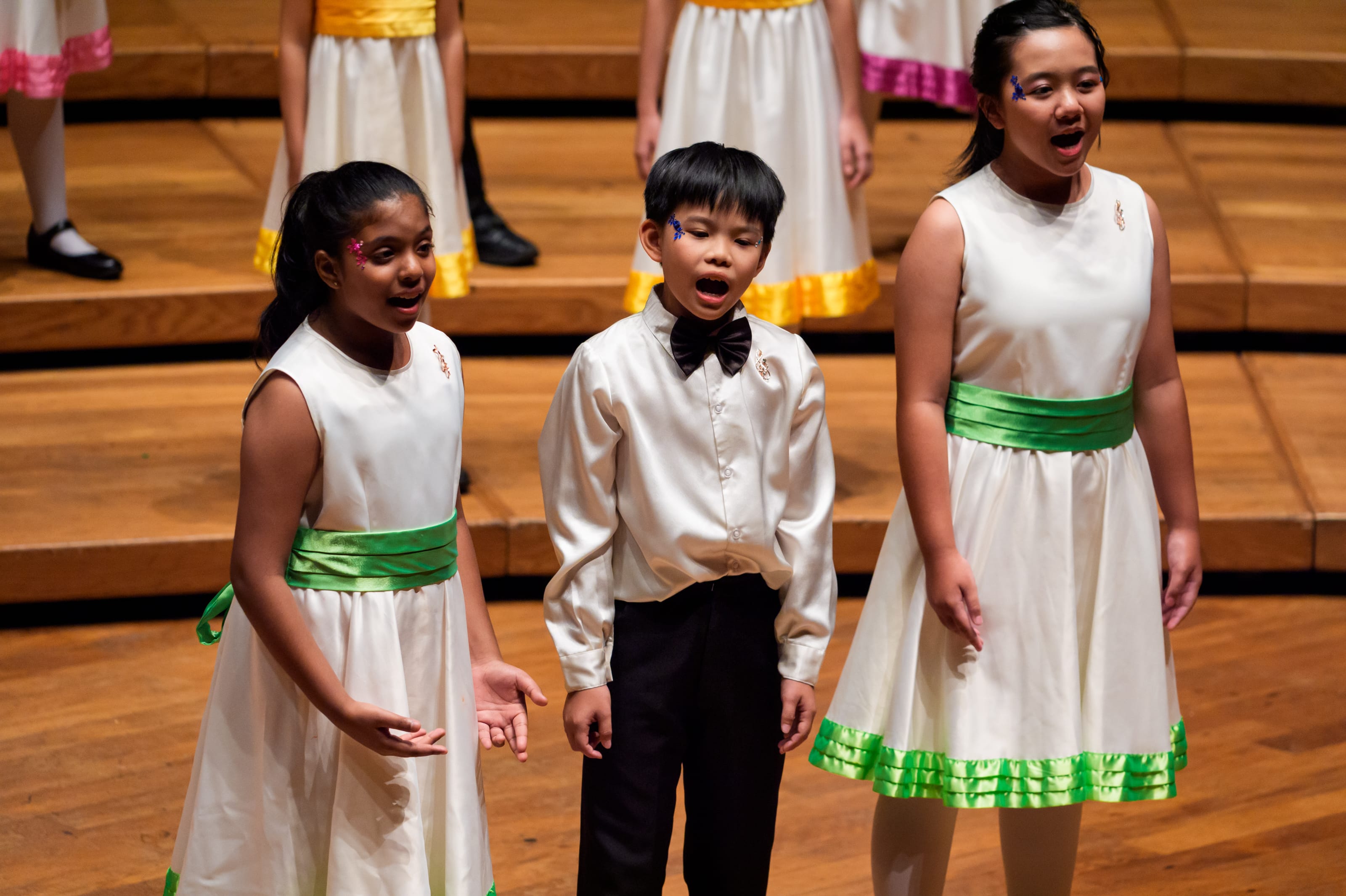 Three children sing onstage; two in white dresses with green sashes, one in white shirt and bow tie.