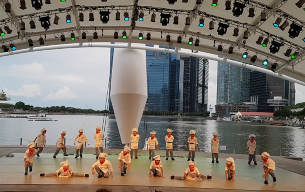 Children in chef costumes perform on an outdoor waterfront stage with city skyscrapers visible in the background.