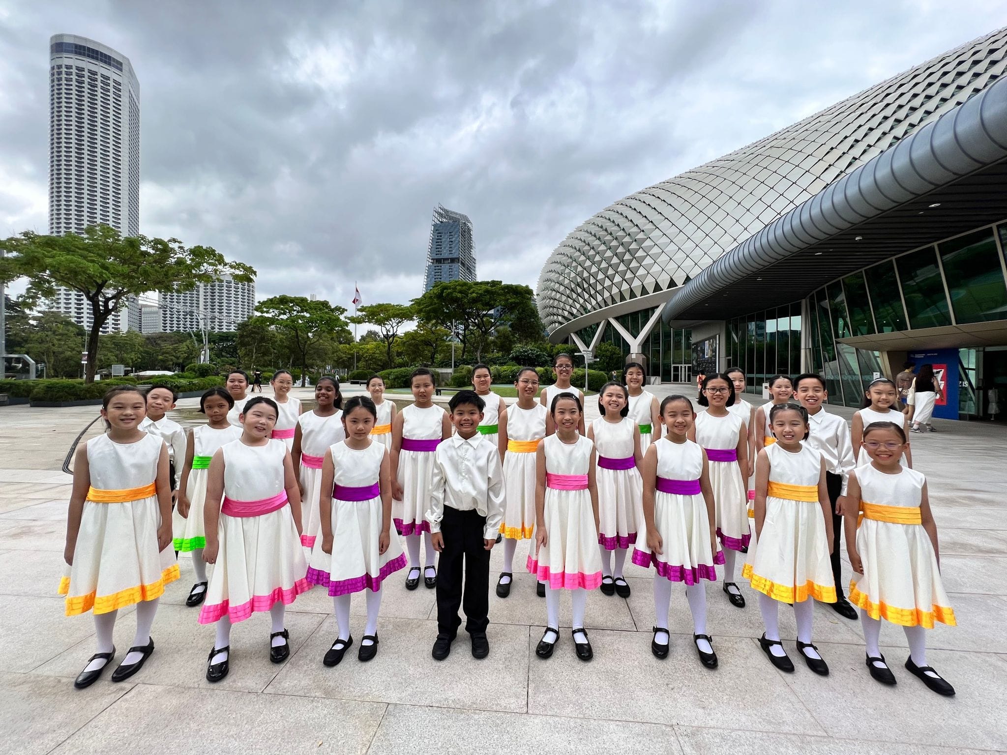 A group of children in white dresses with colorful sashes pose outdoors near Singapore's Esplanade building.