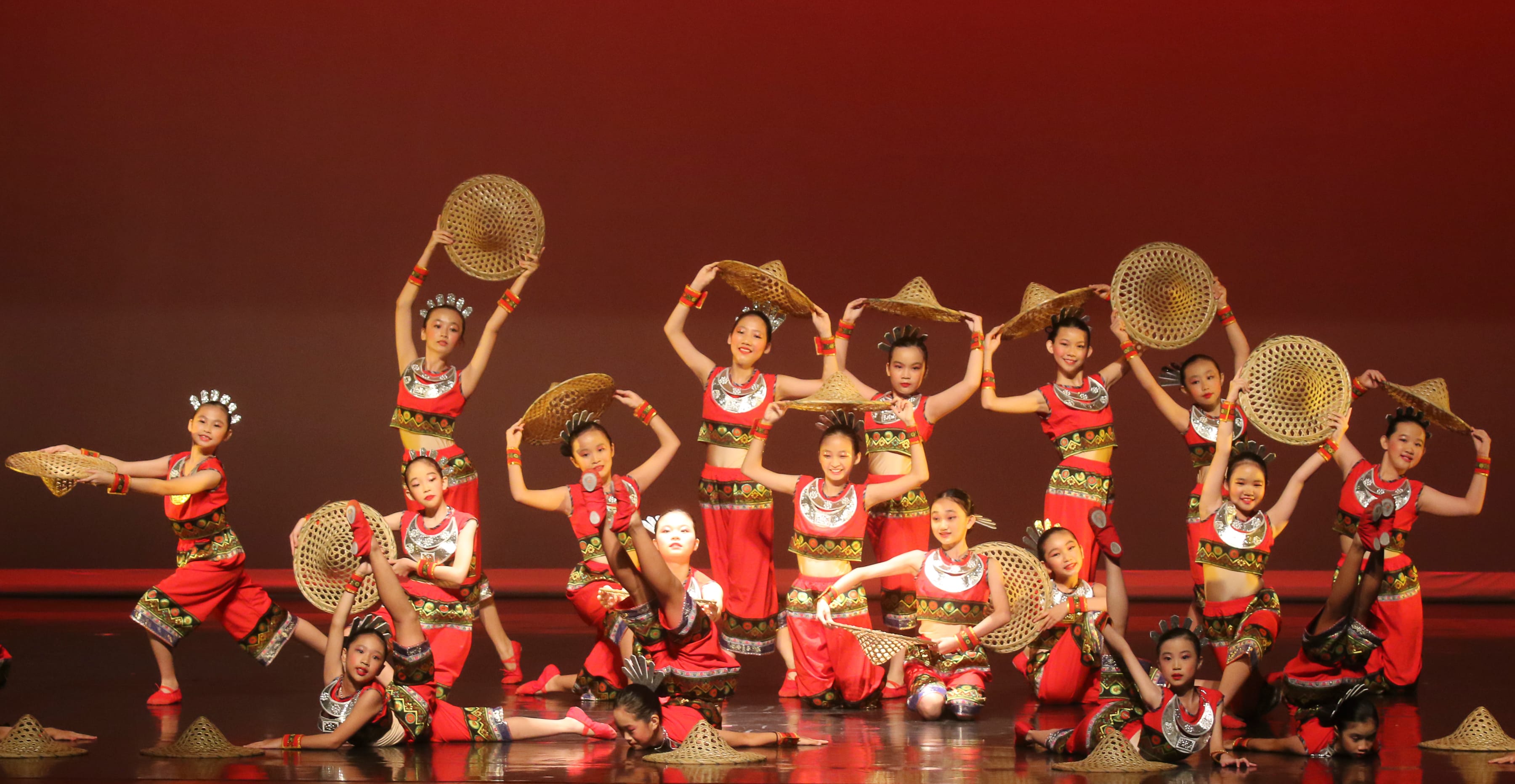 Children in red traditional Chinese costumes perform on stage, holding woven bamboo hats against a red backdrop.