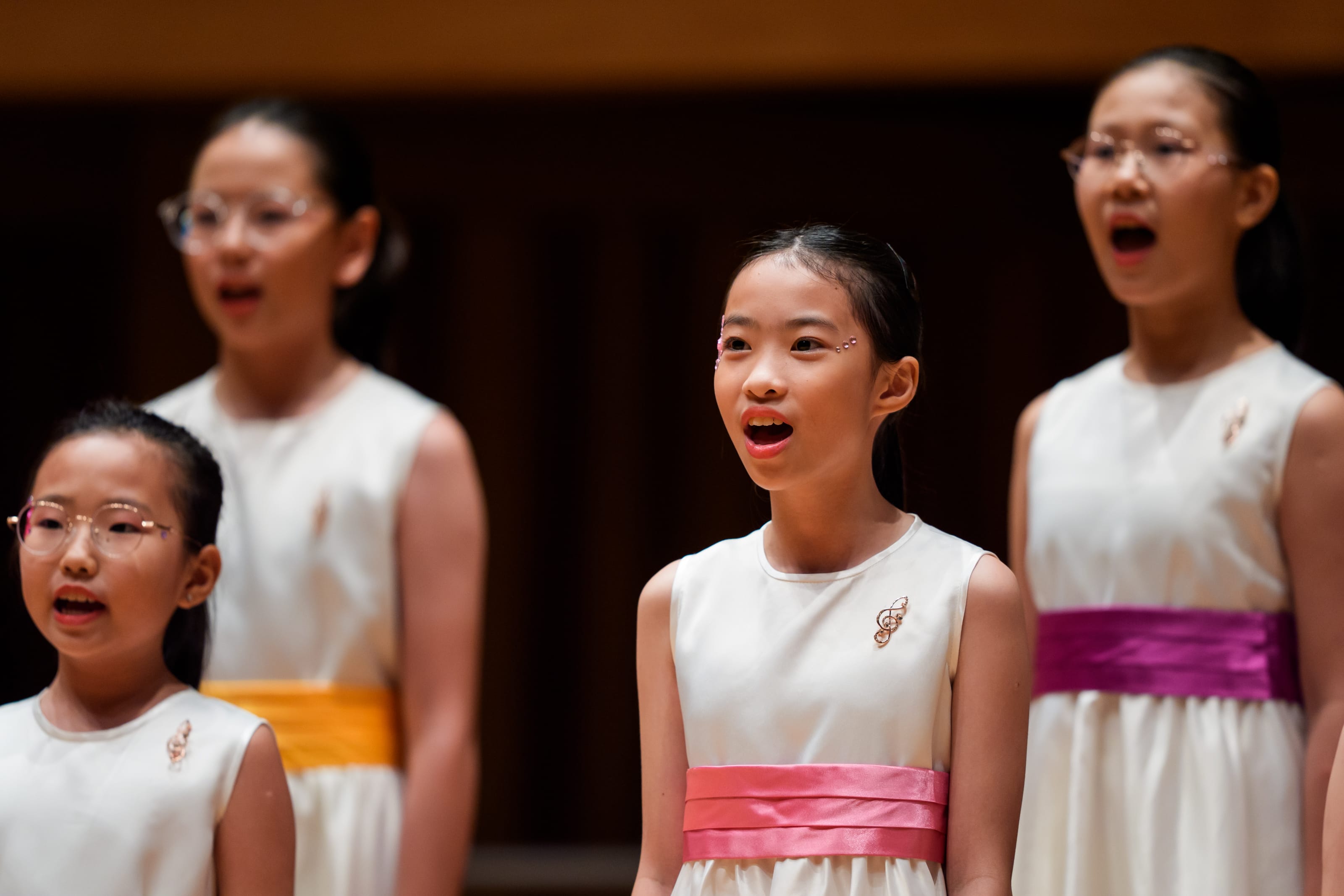Four young girls in white dresses with colorful sashes sing open-mouthed during a choir performance on stage.