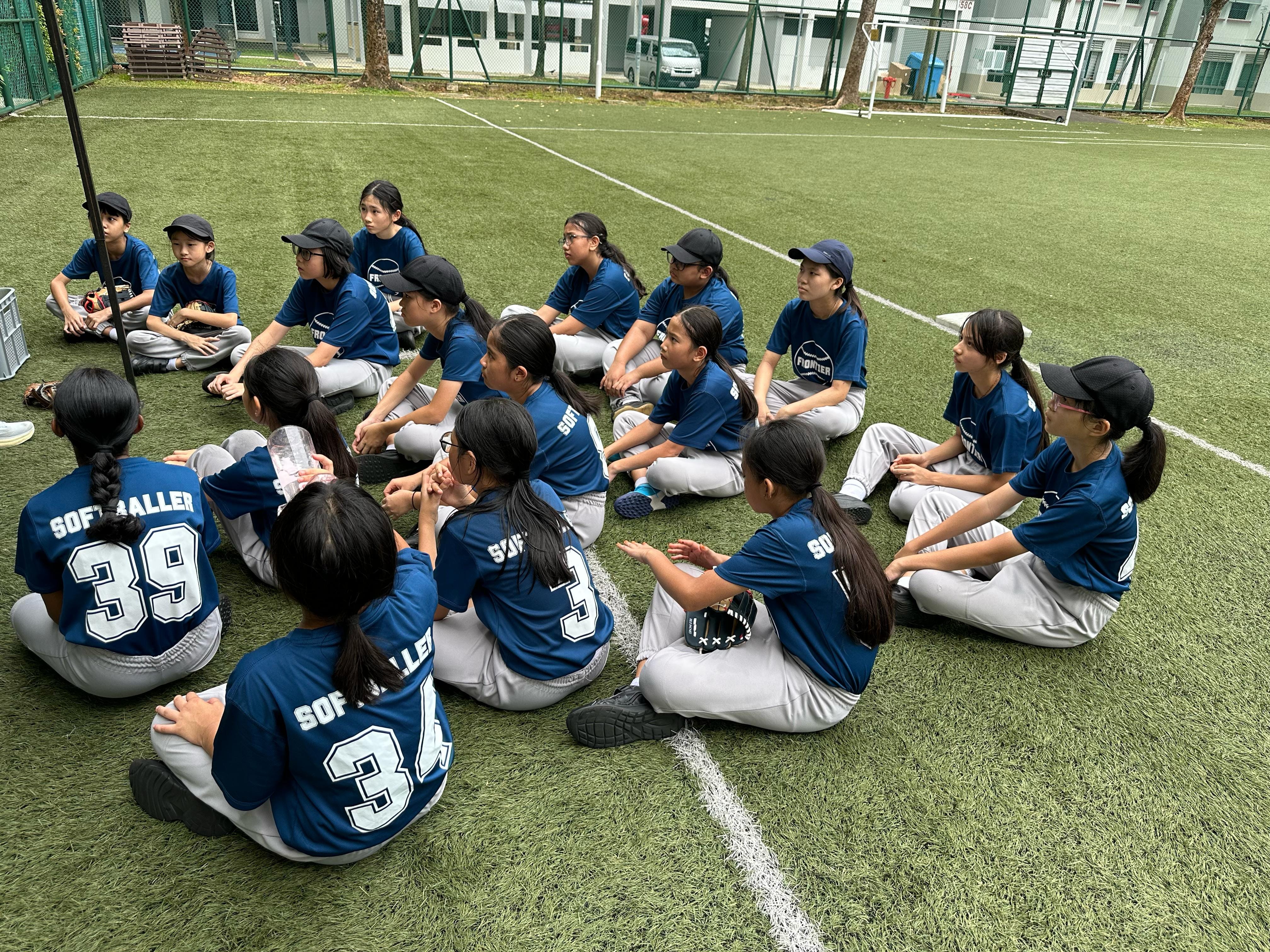 A youth softball team in blue jerseys and caps sits in a circle on a green turf field.