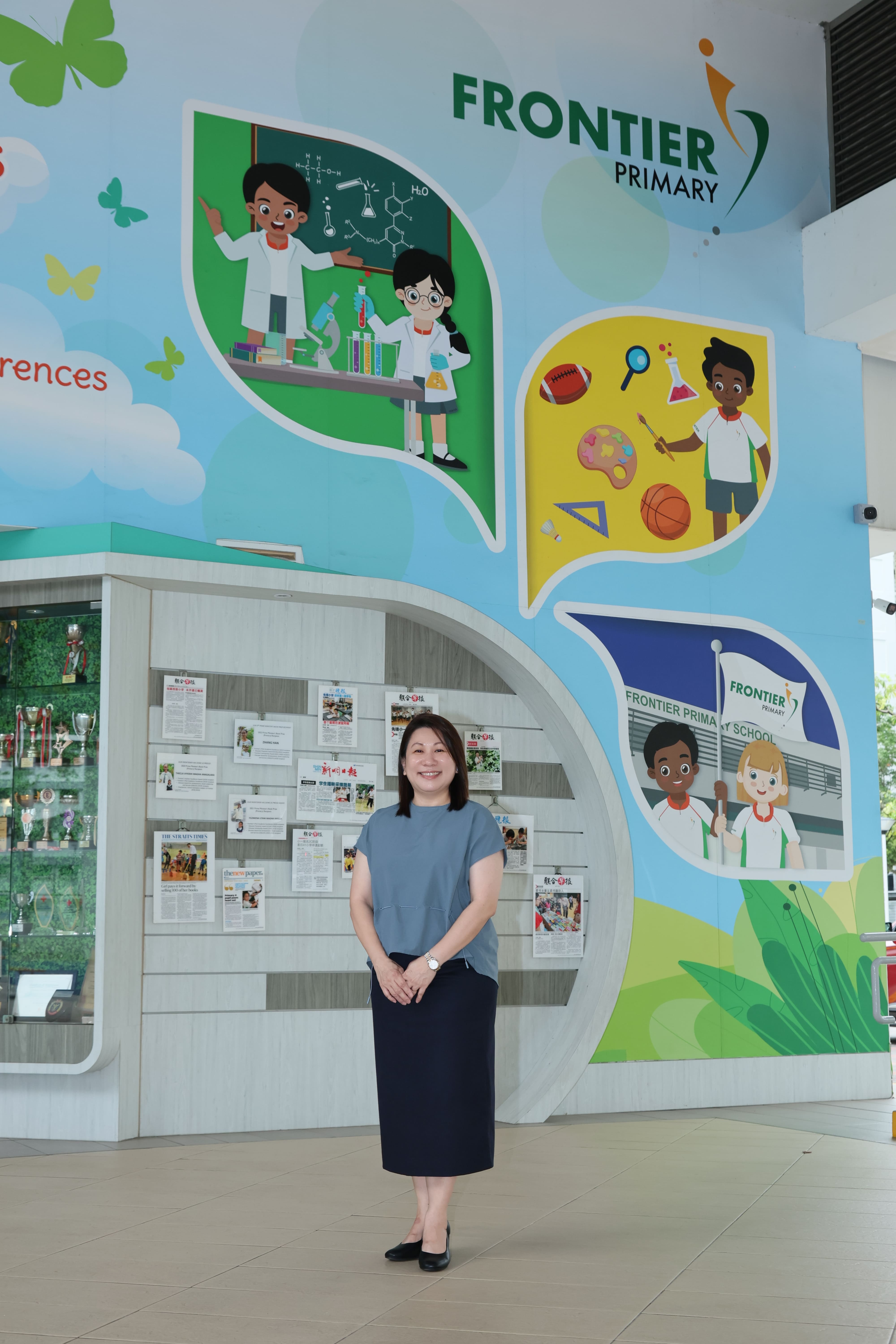 Woman in grey top and navy skirt stands smiling before a colorful Frontier Primary School mural.