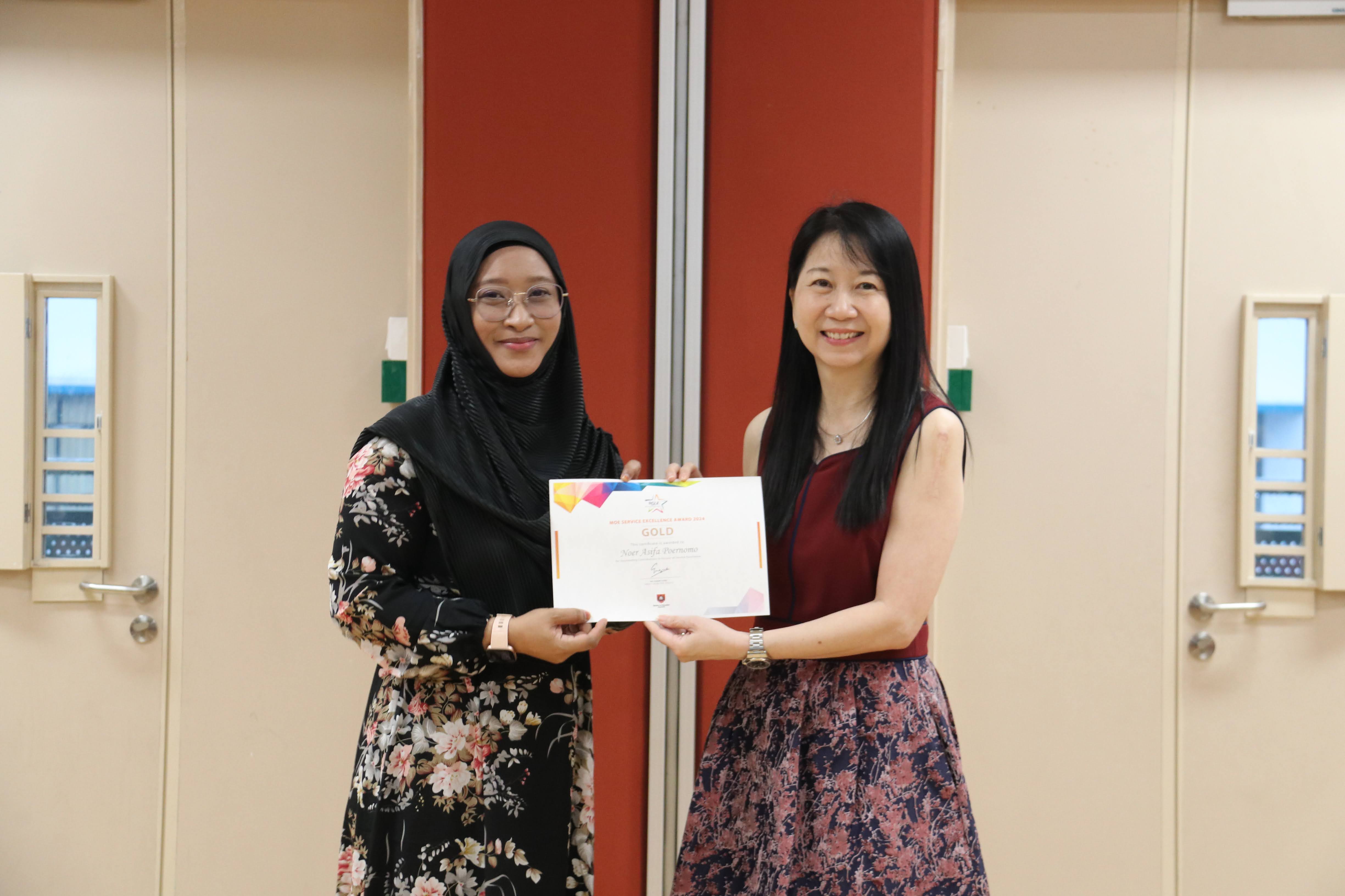 Two women smile while holding a colorful Gold award certificate together indoors.