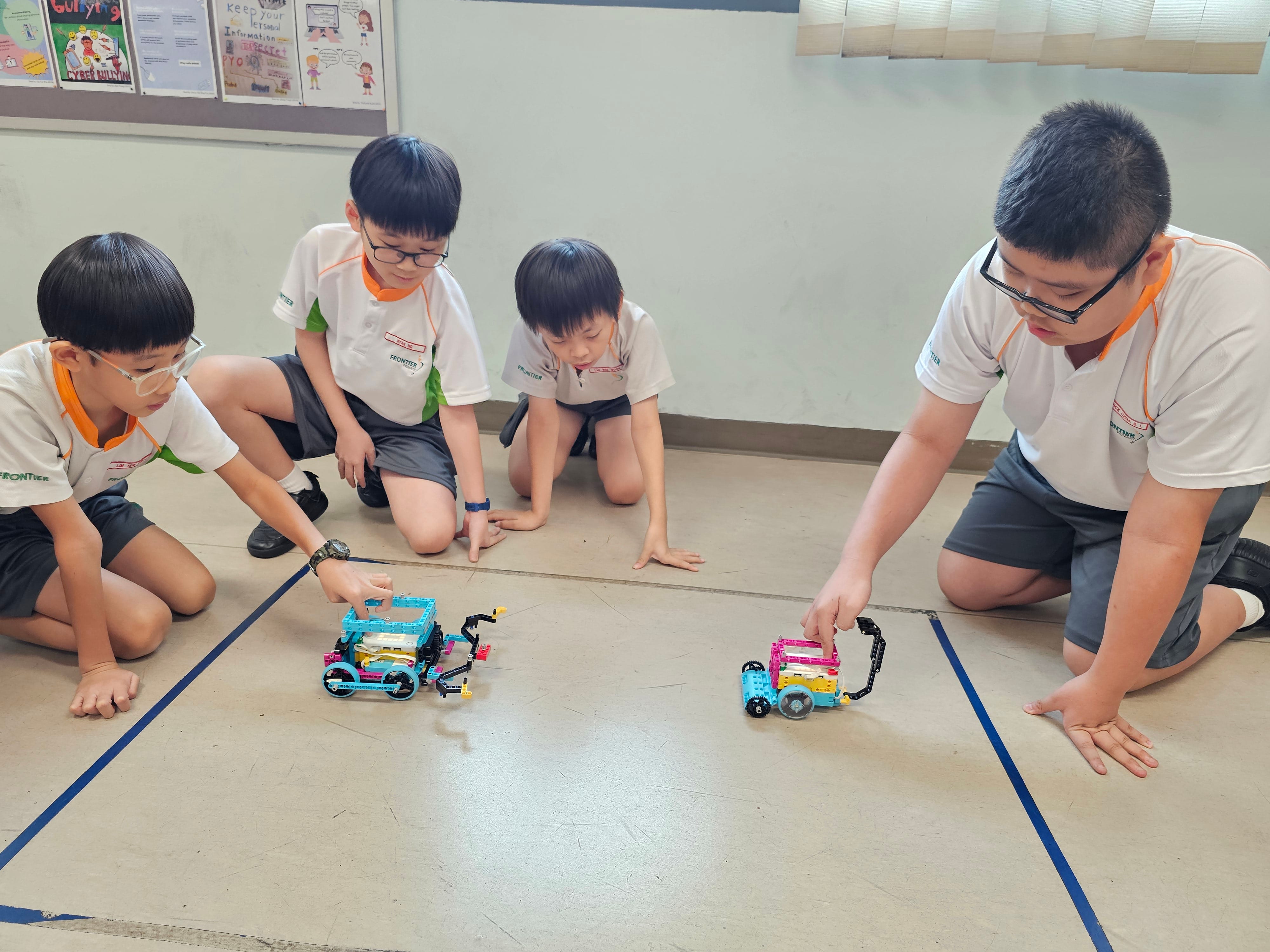 Four school boys in uniforms kneel on a floor, operating two small colorful LEGO robots within taped boundary lines.