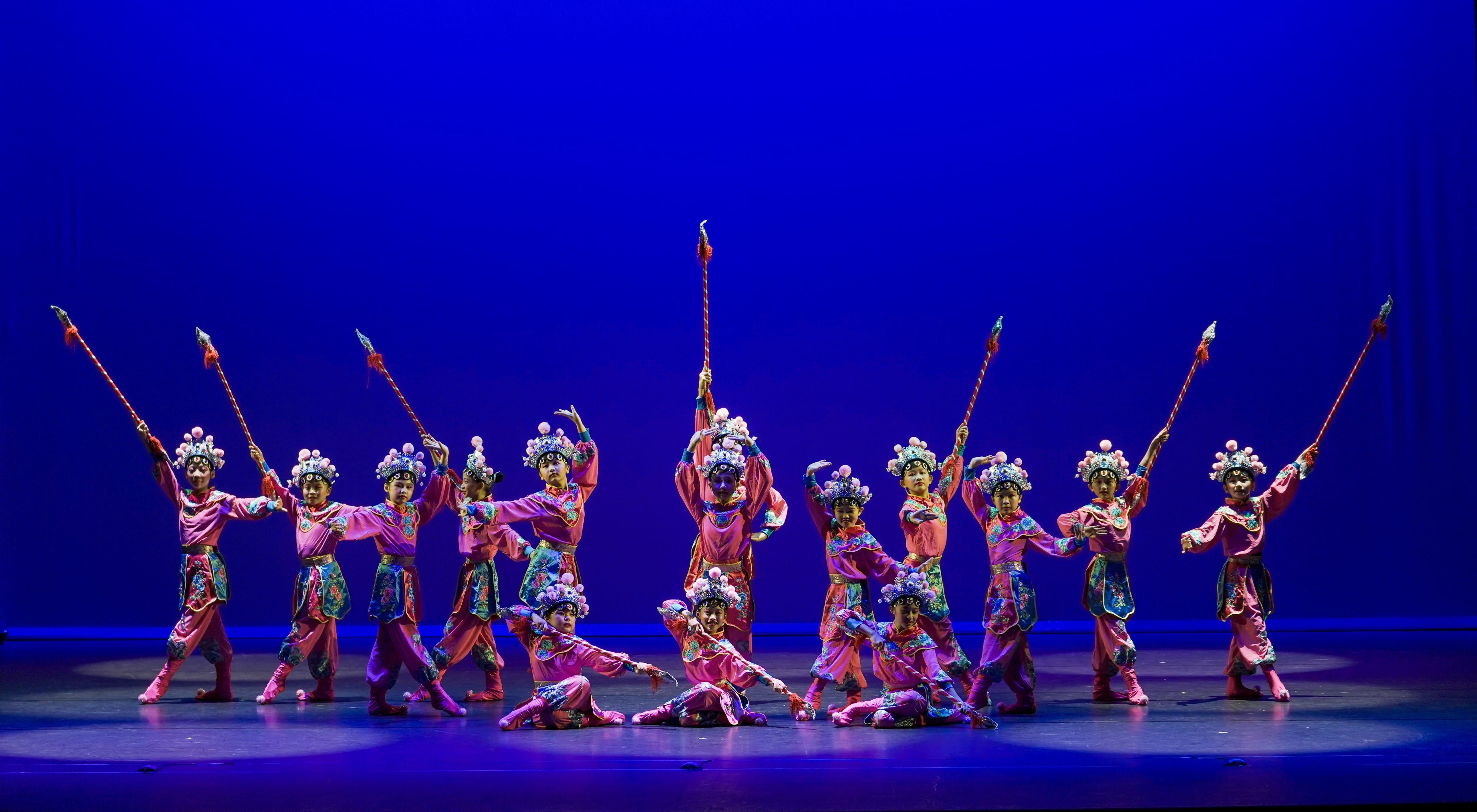 Performers in pink traditional Chinese opera costumes and headdresses hold spears on a blue-lit stage.