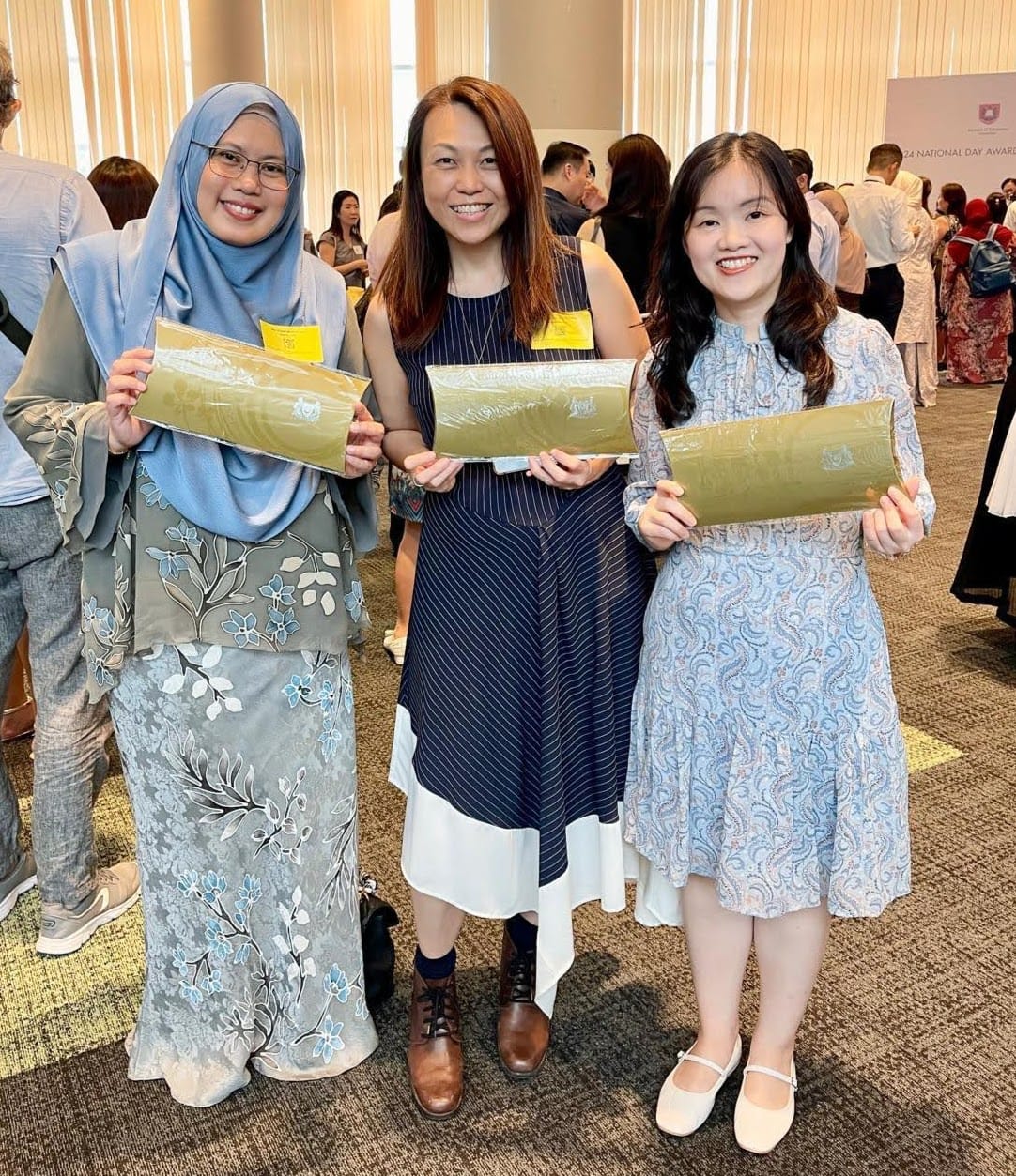 Three smiling women hold green wrapped packages at a National Day Awards ceremony in a crowded hall.