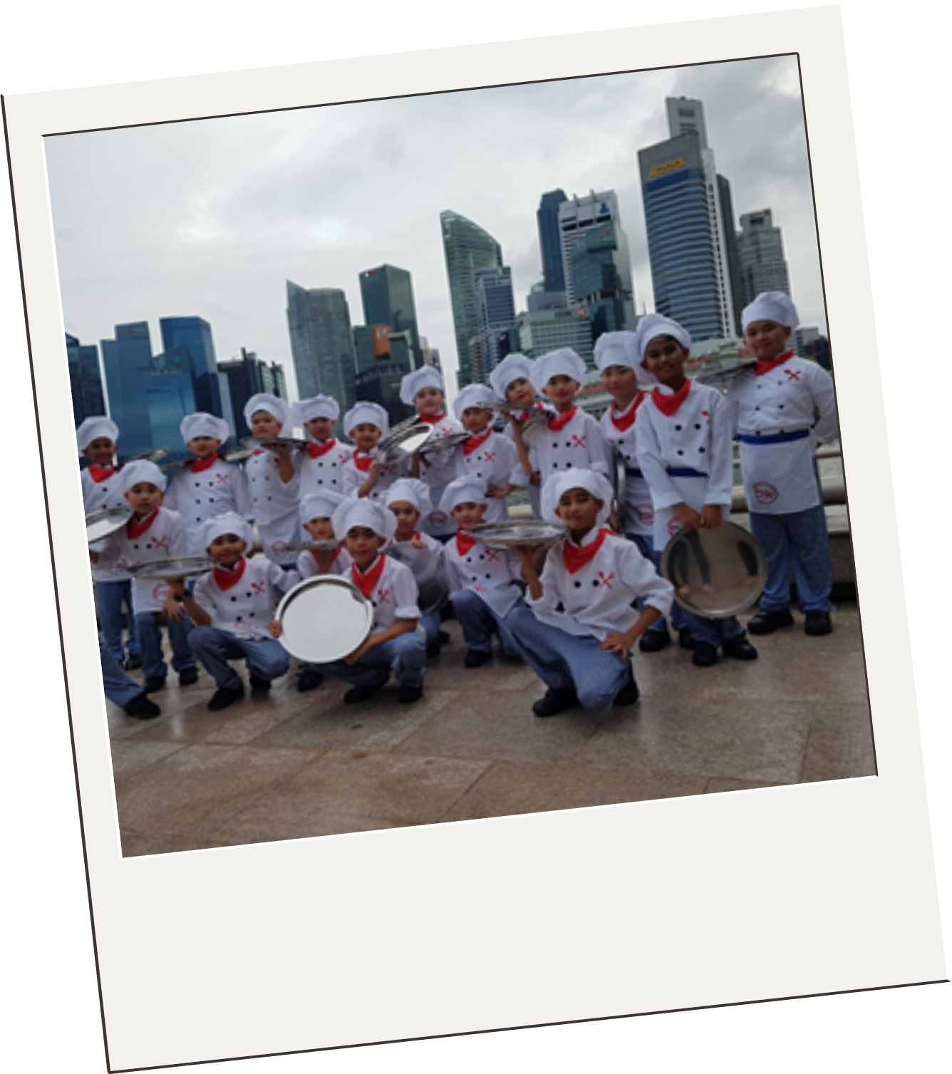 A group of children in white chef uniforms and hats holding silver trays, posing in front of a city skyline.