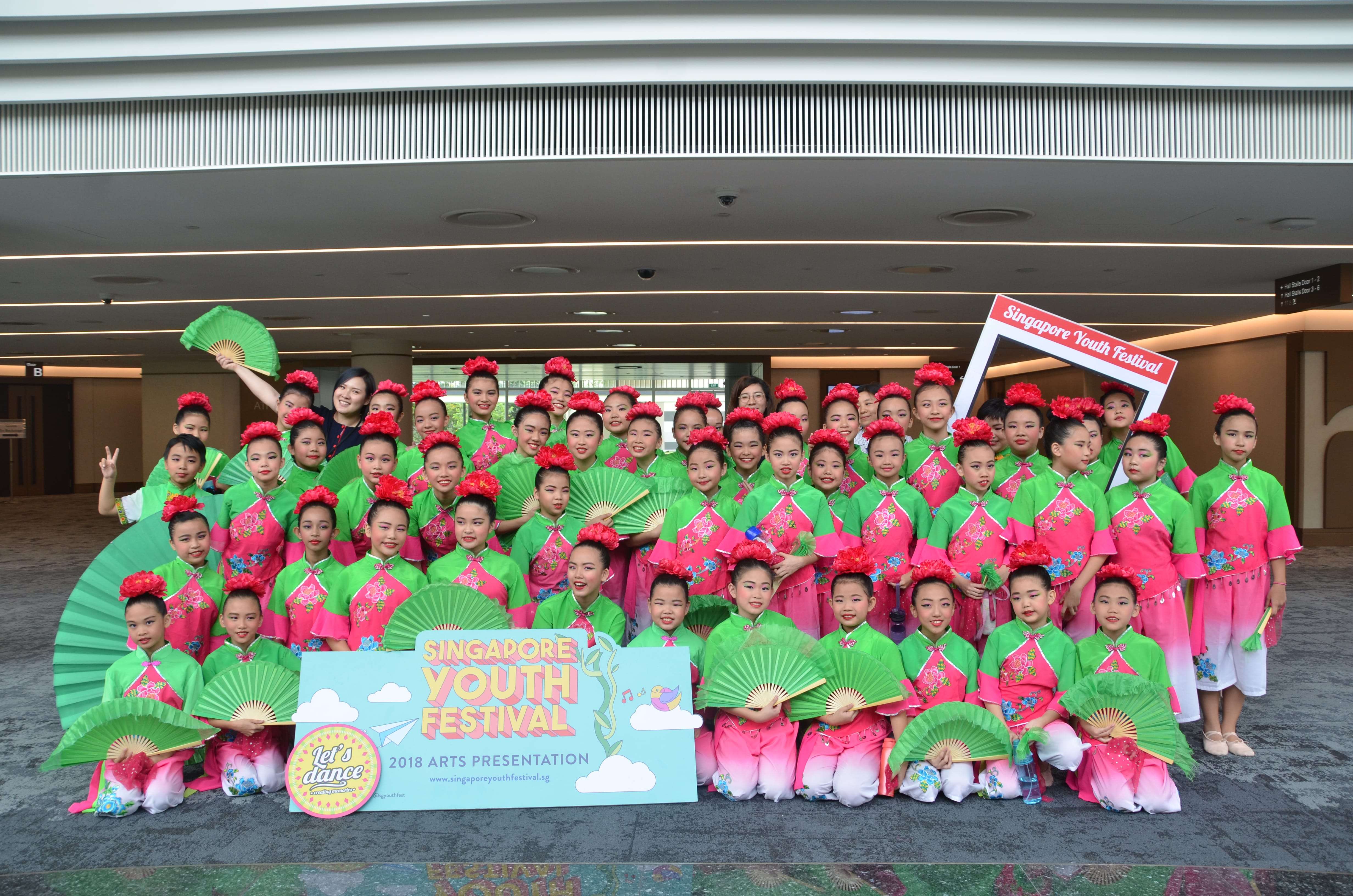 Group of children in green and pink costumes holding green fans, posing with Singapore Youth Festival sign