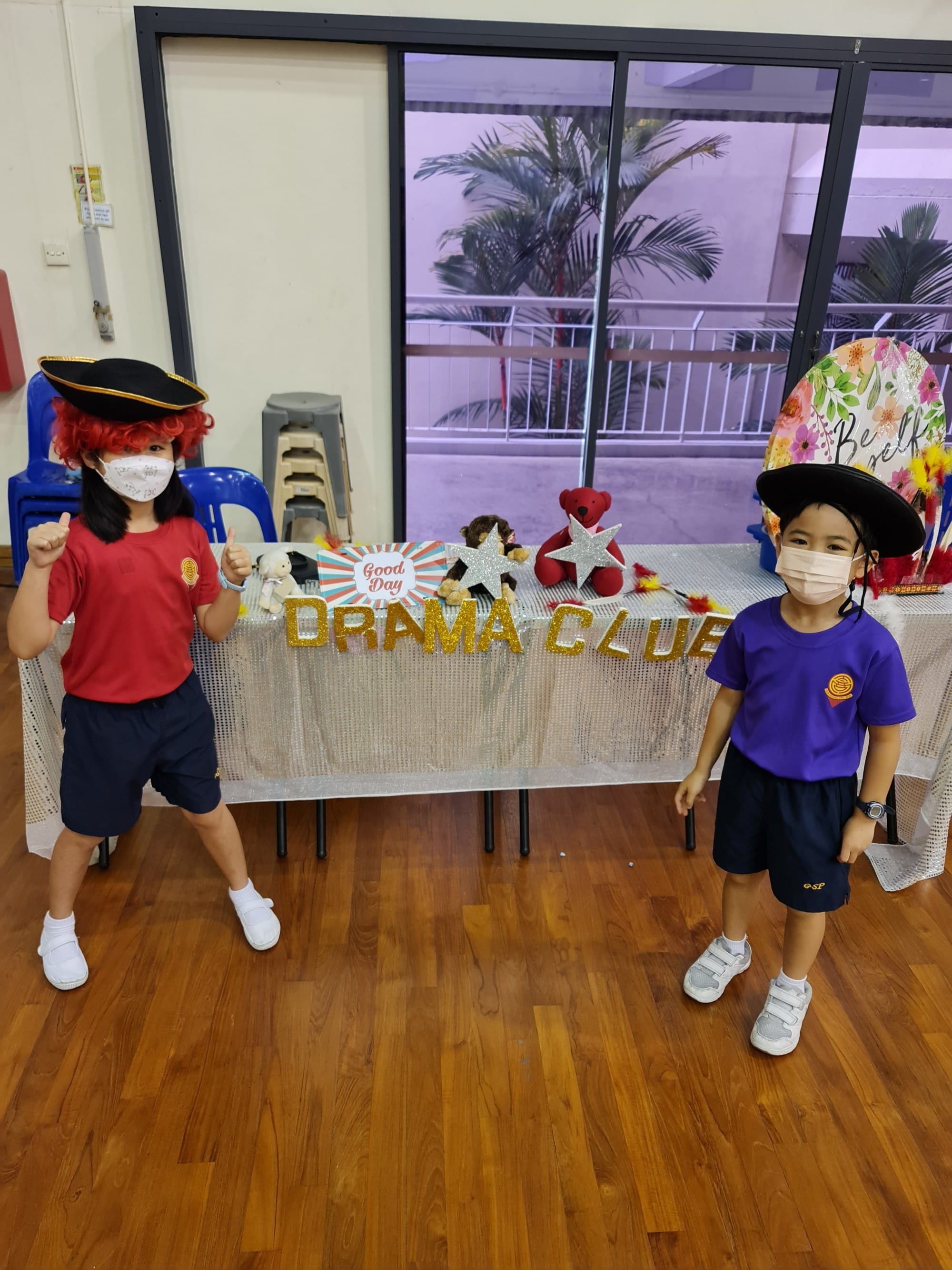 Two masked children in costumes with hats pose in front of a Drama Club table with gold letters and star decorations.
