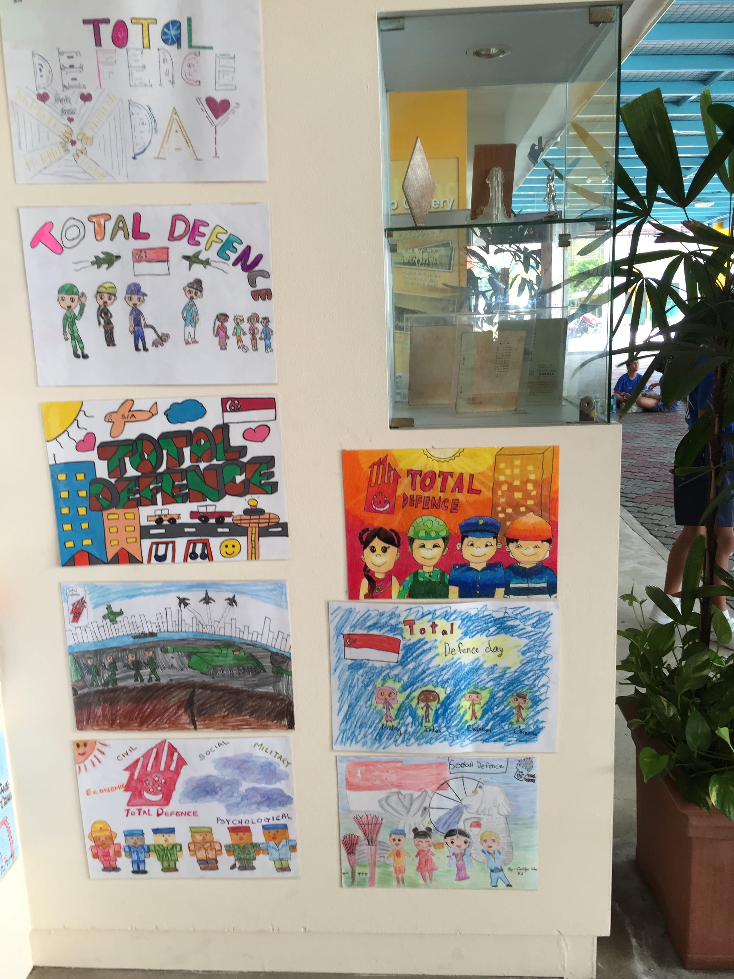 Seven colorful children's drawings about Total Defence Day displayed on a white wall near a glass entrance.
