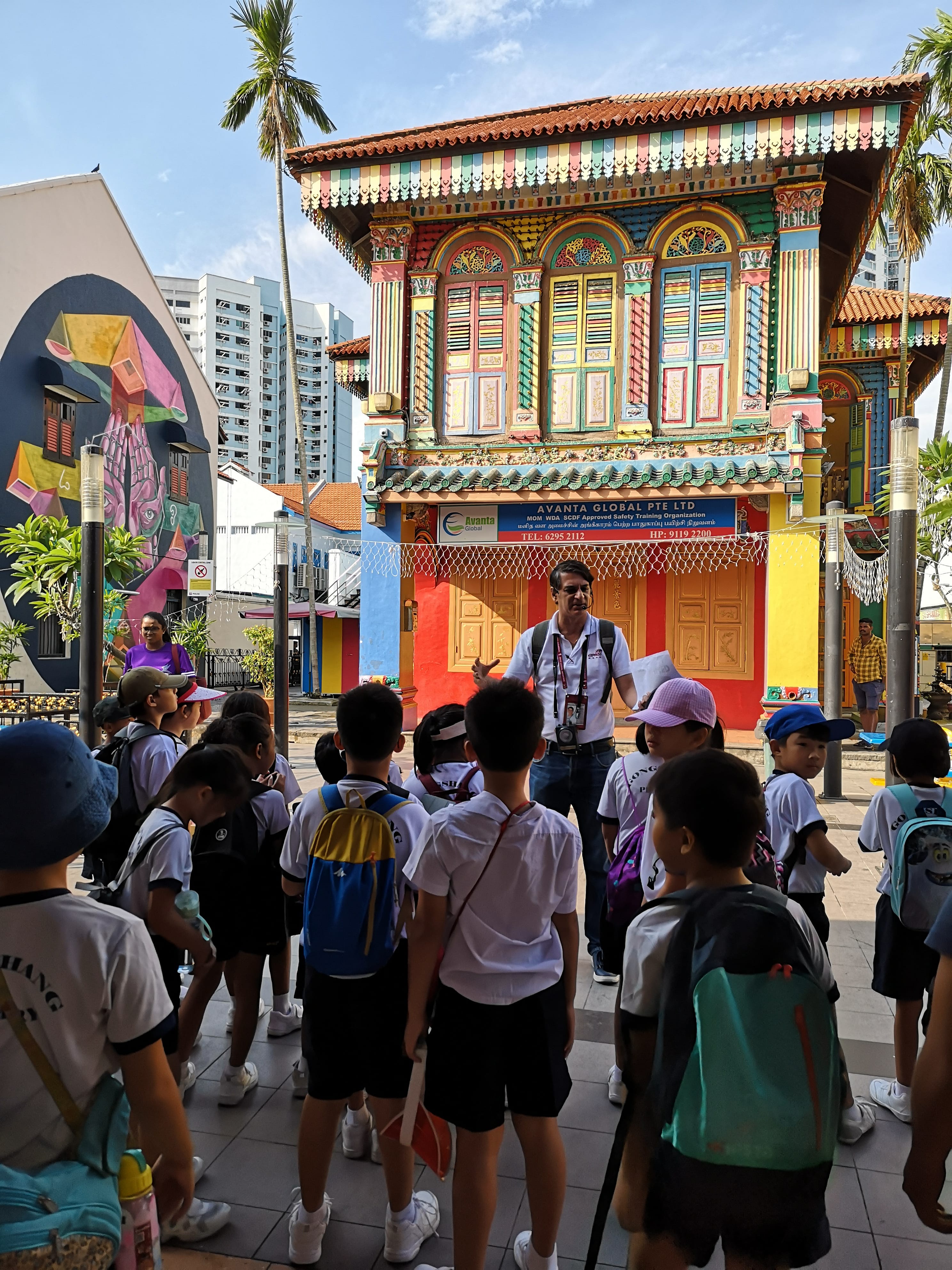 Tour guide speaks to a group of schoolchildren in front of a colorful ornate building on a sunny street.