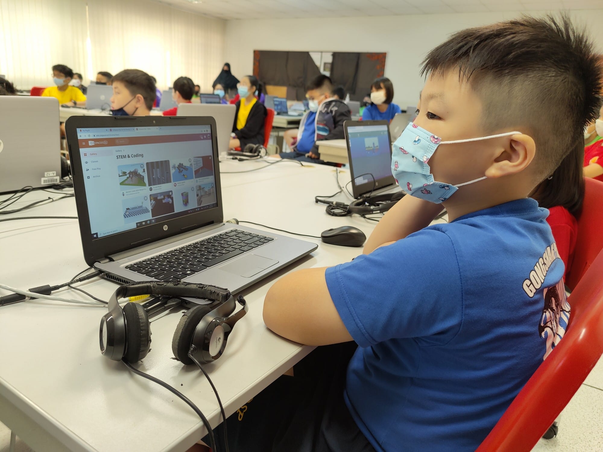 Young masked boy in blue shirt uses a laptop showing a STEM and Coding page in a classroom with other masked children.