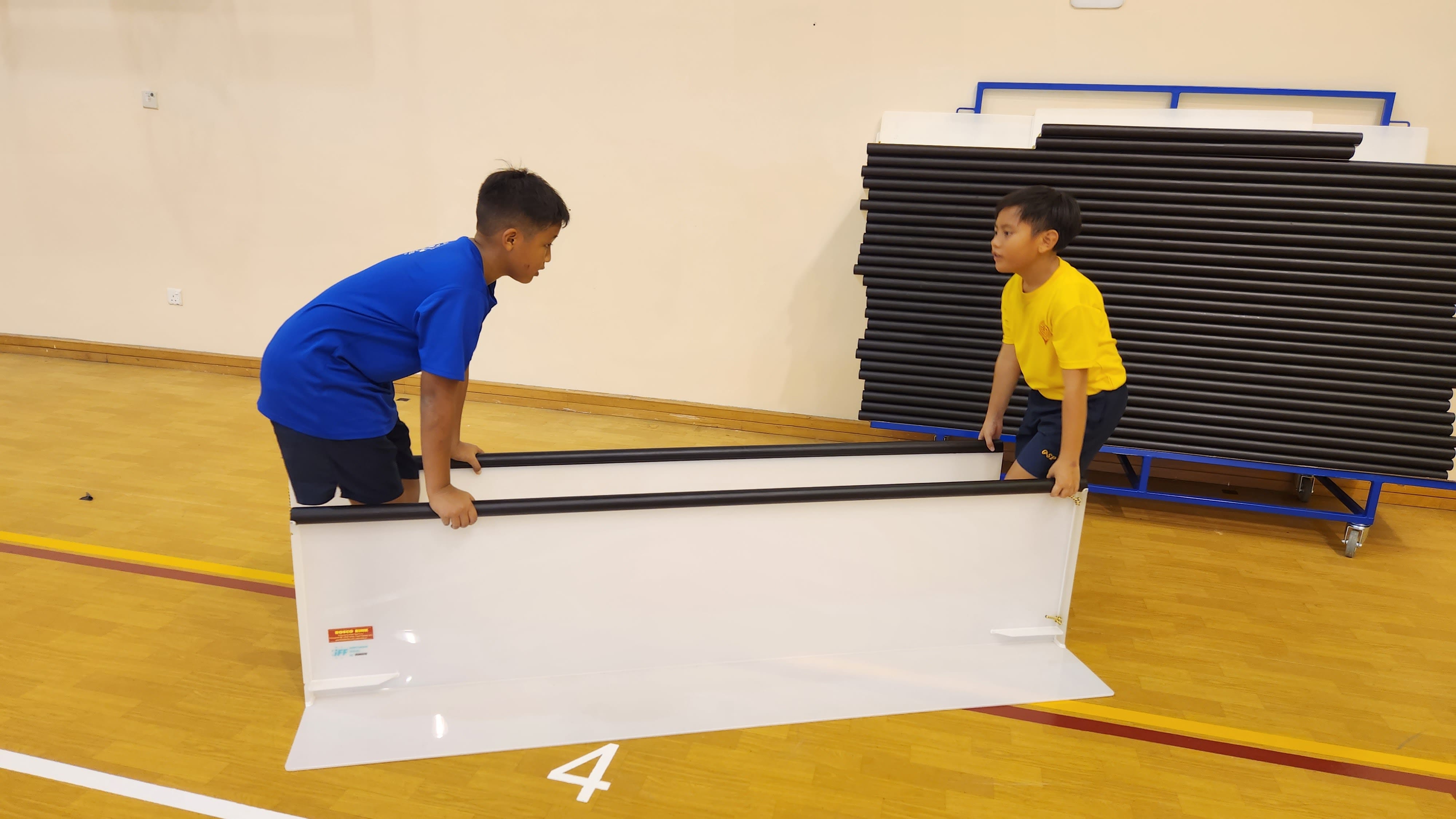 Two boys carry a large white panel together on a gymnasium floor near stacked exercise mats.