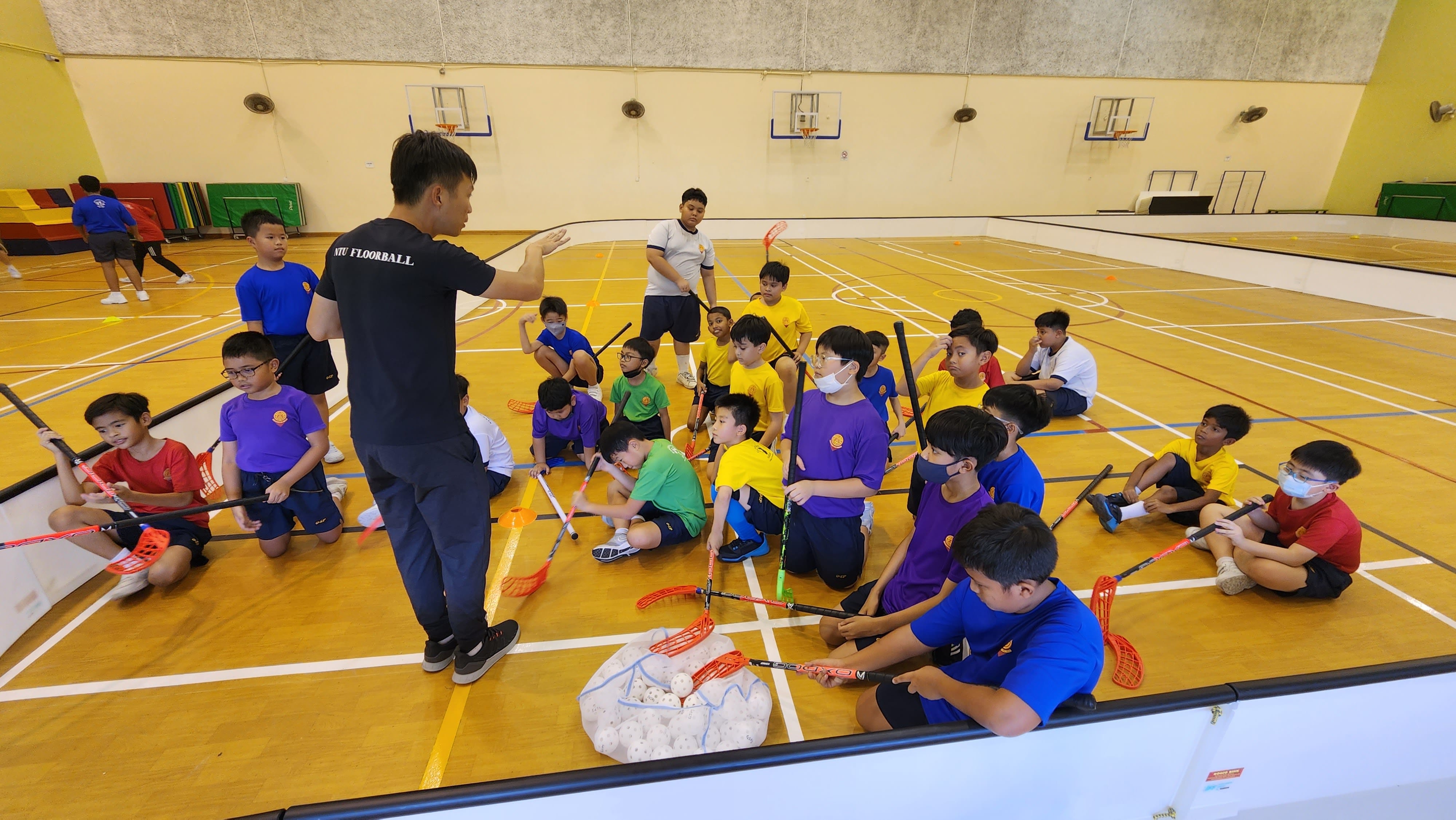 A coach instructs a group of children holding floorball sticks seated on an indoor sports court.