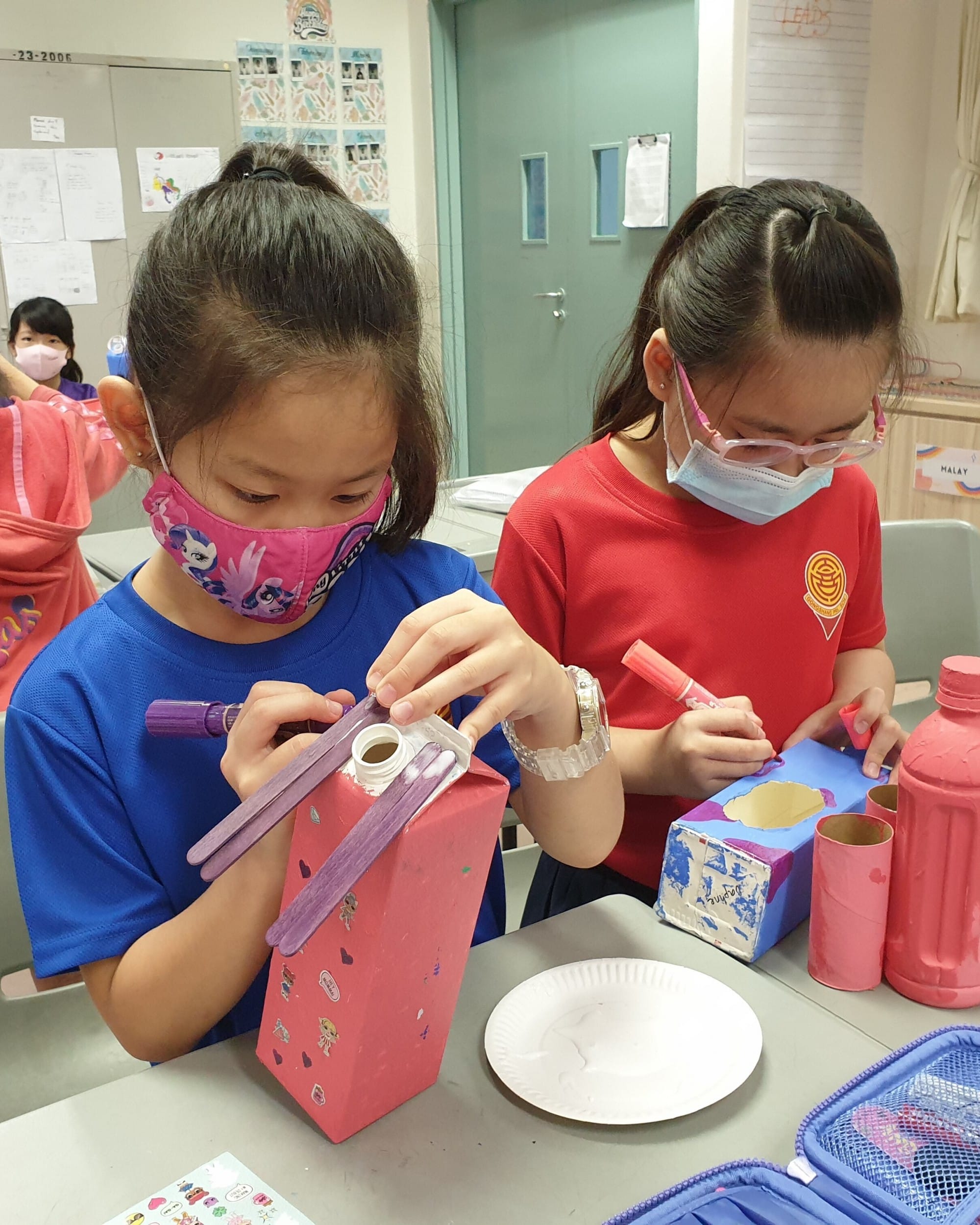 Two masked girls decorate painted cardboard boxes with markers at a classroom table.