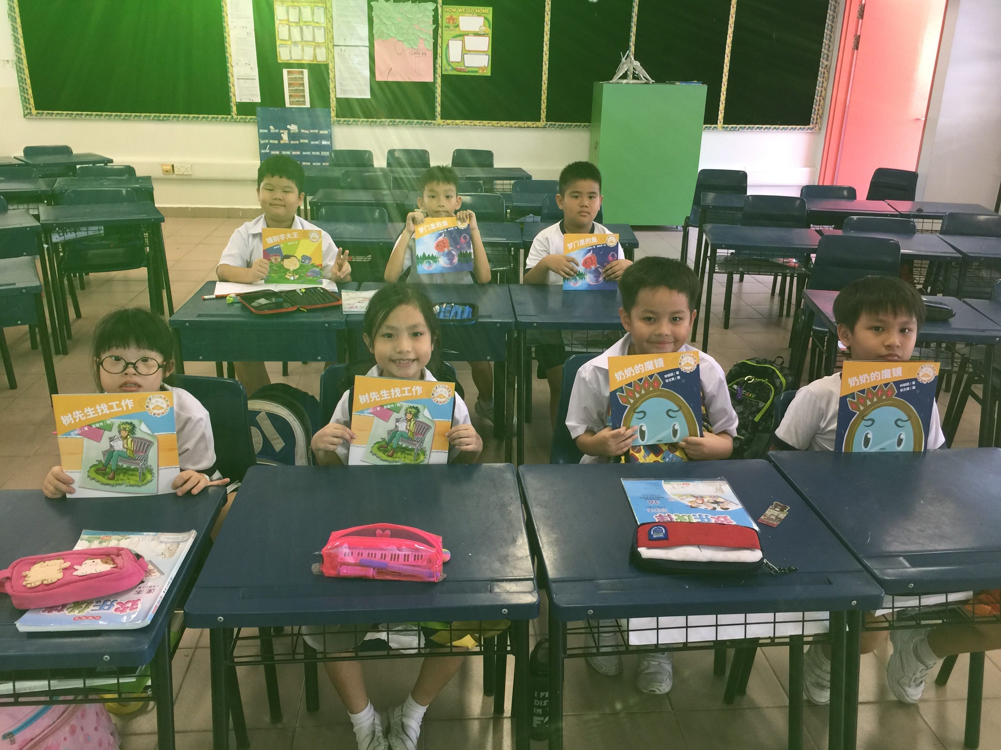 Seven young students in white uniforms sit at school desks holding up colorful Chinese-language picture books.