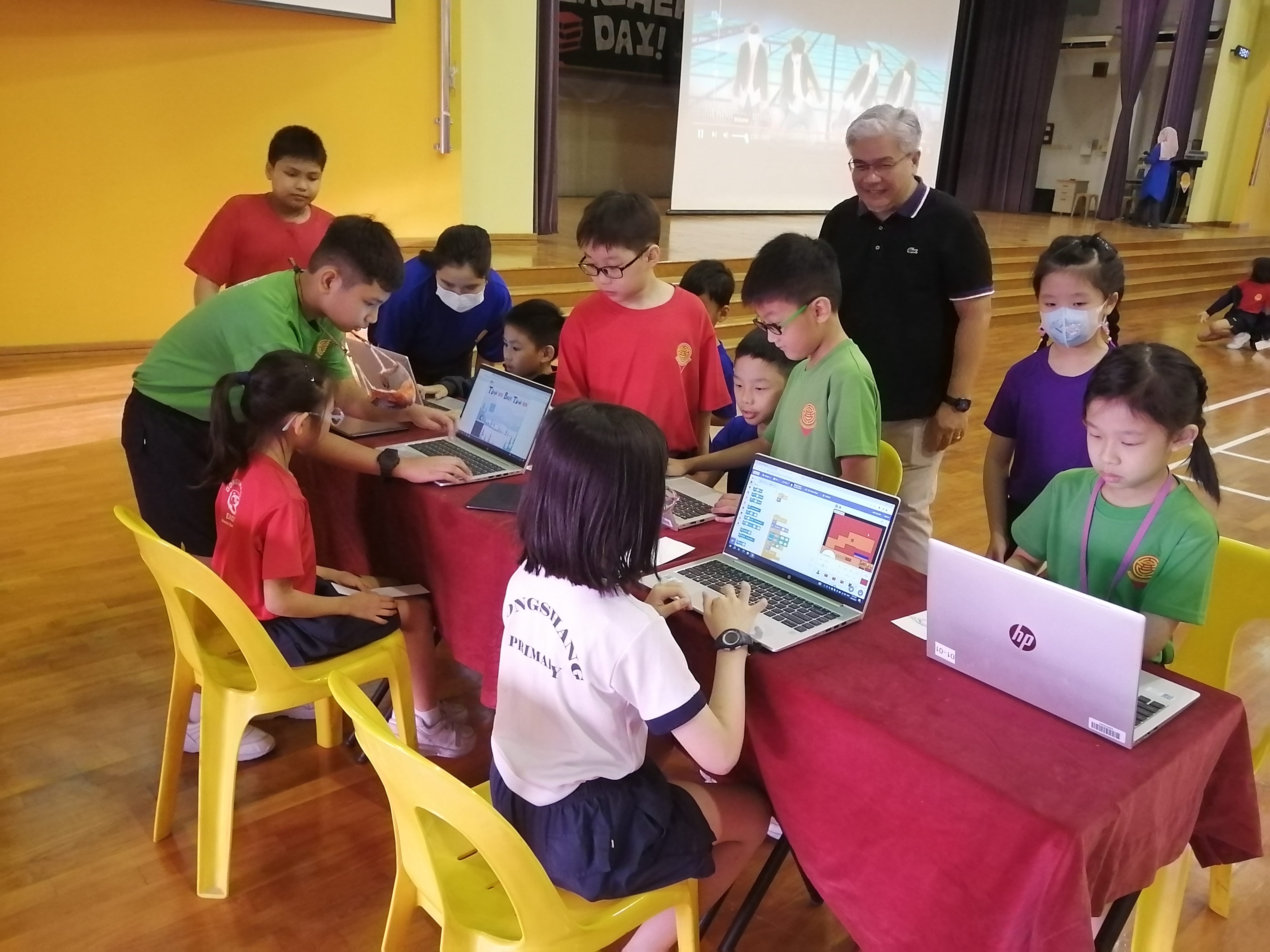 Children gather around laptops at a red-covered table in a school hall while a teacher watches.