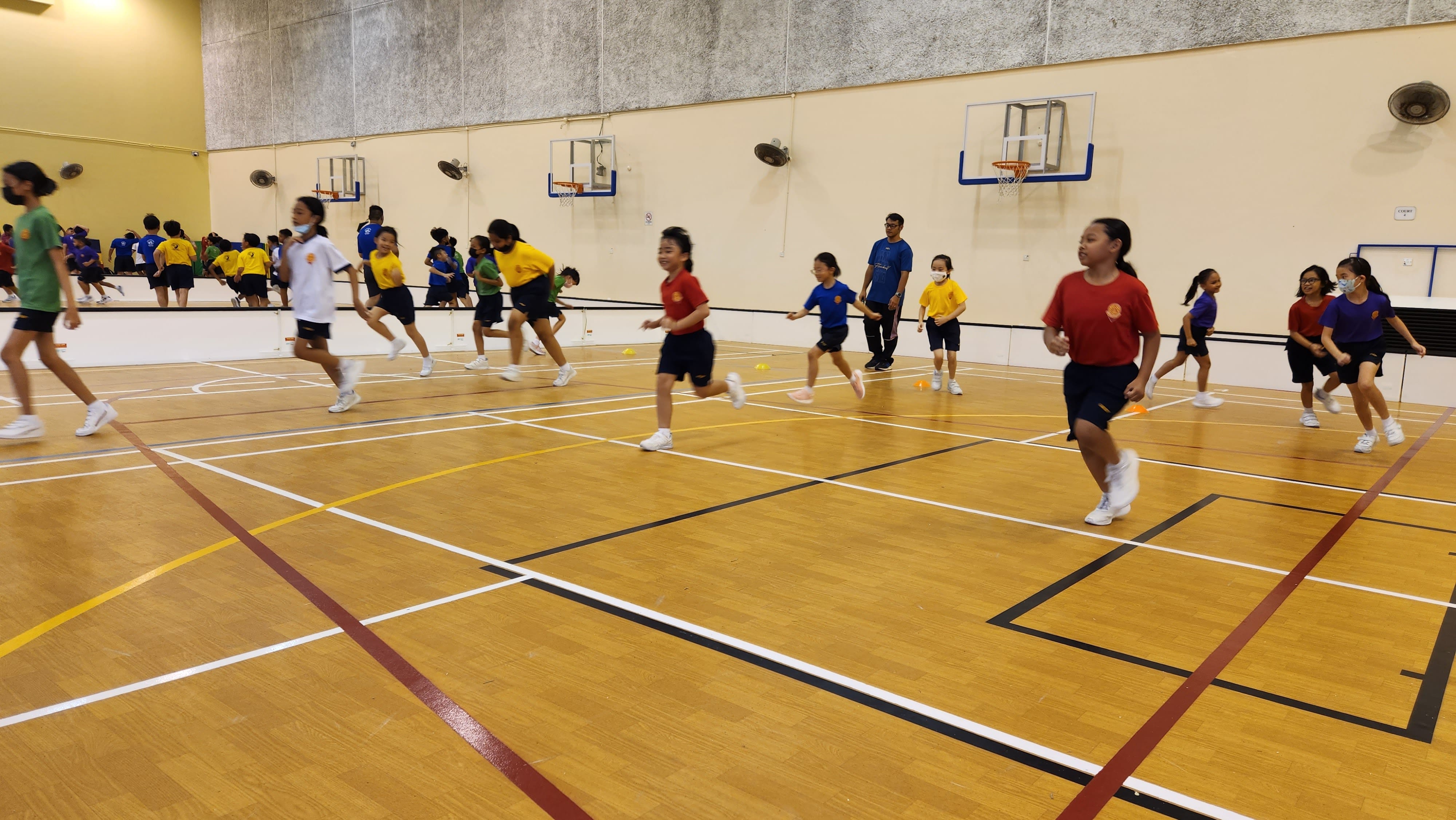 Children in colorful uniforms run across an indoor gymnasium with basketball hoops on the walls.