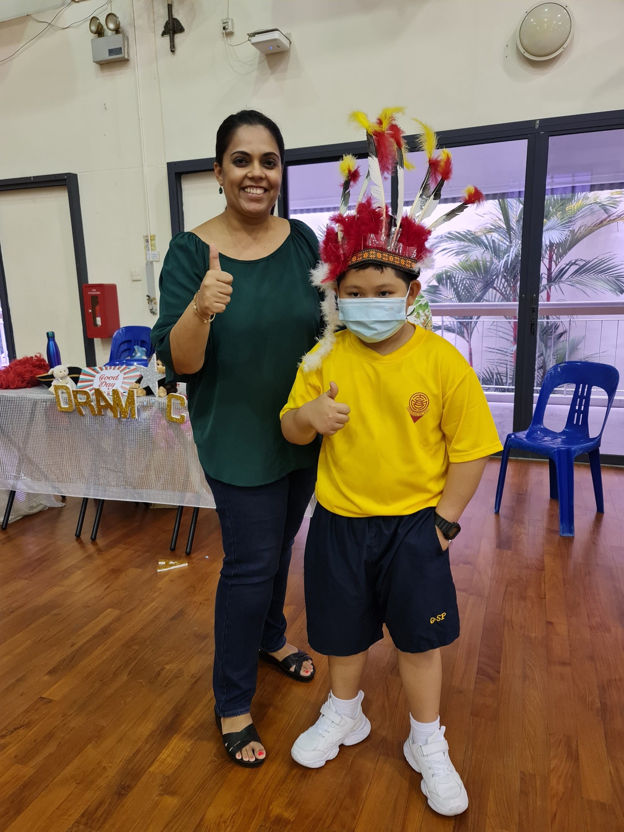 A woman in green and a masked boy in yellow wearing a feathered headdress both give thumbs up indoors.