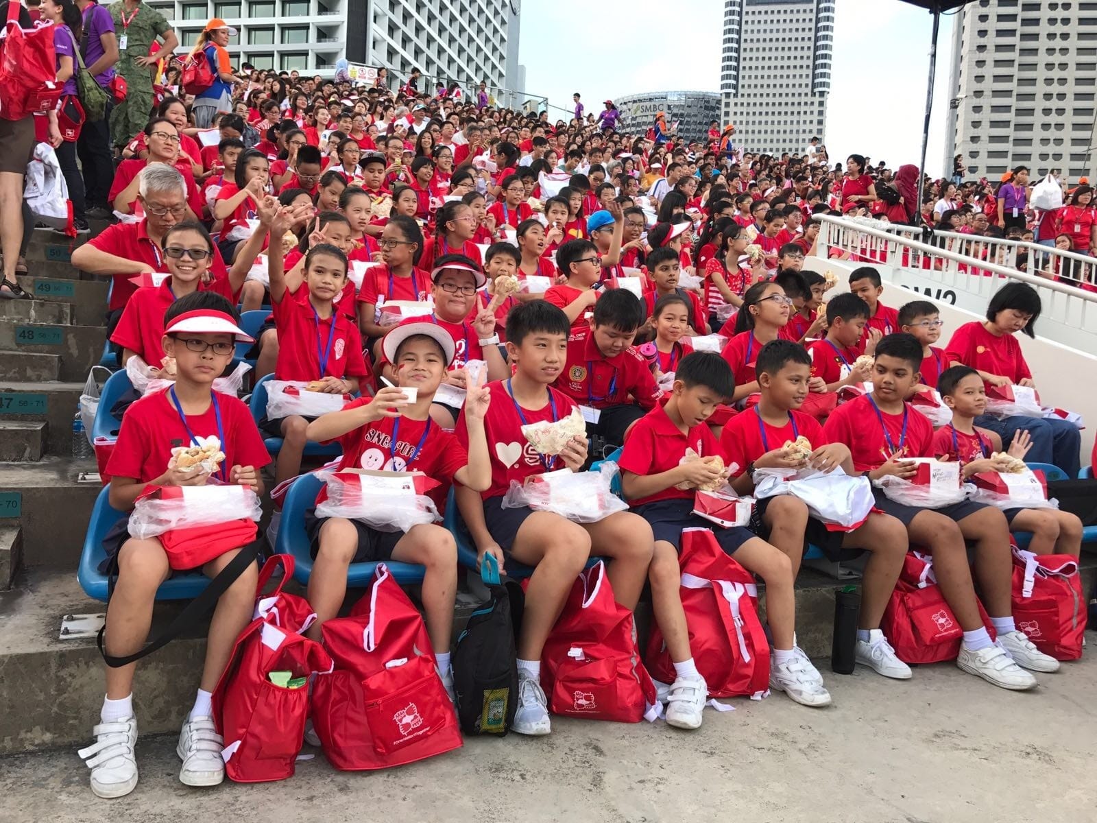 Large crowd of children in red shirts seated on stadium bleachers, holding food and red bags at an outdoor event.