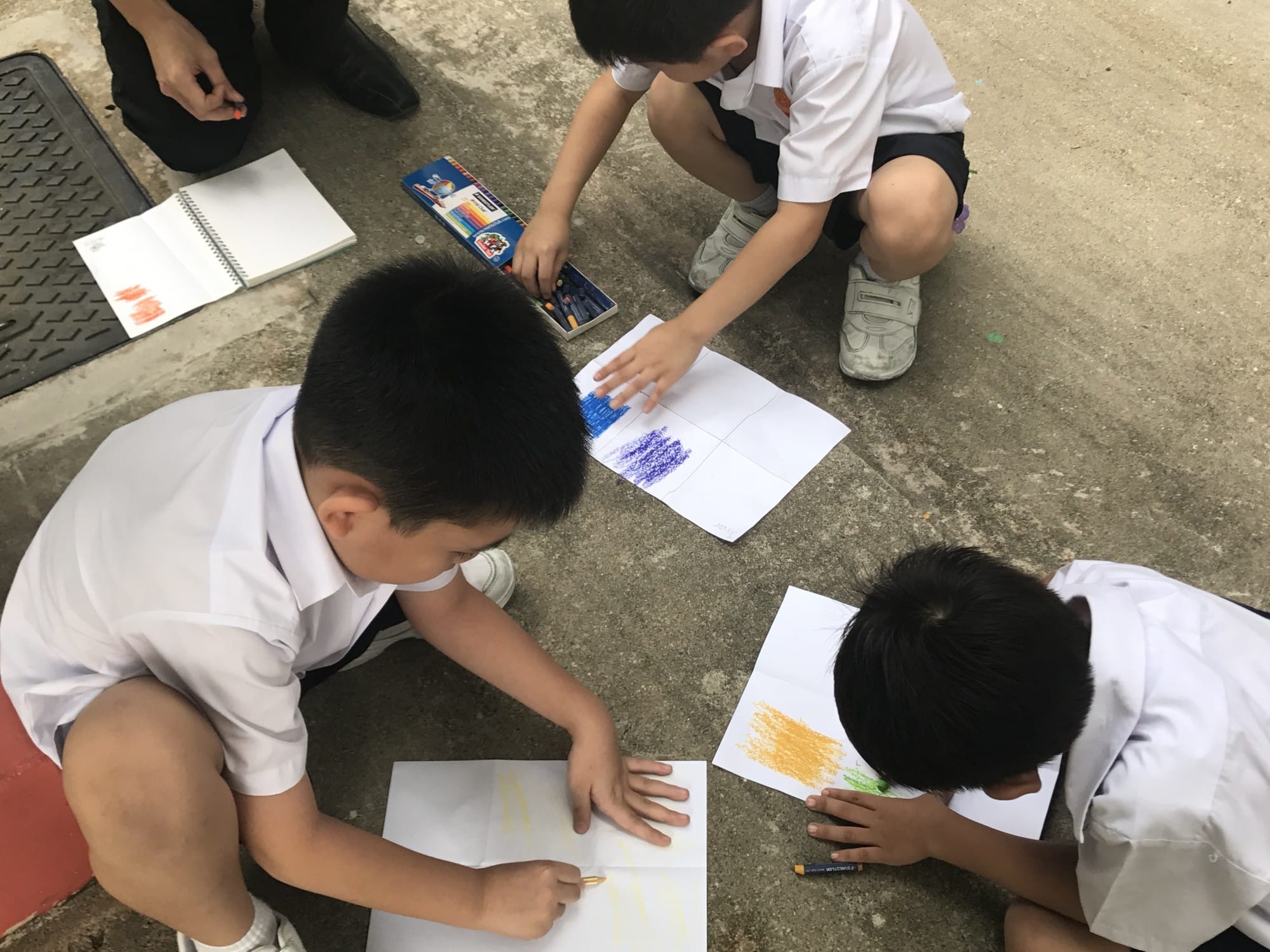Three young boys in white uniforms sit on concrete, drawing with crayons on white paper sheets.