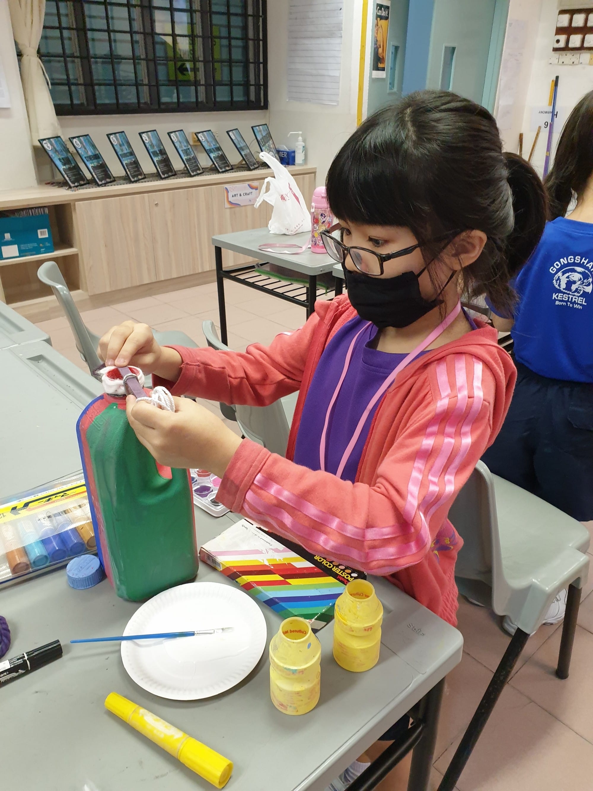 A young girl wearing a black mask and glasses paints a green bottle at a craft table with paints and brushes.