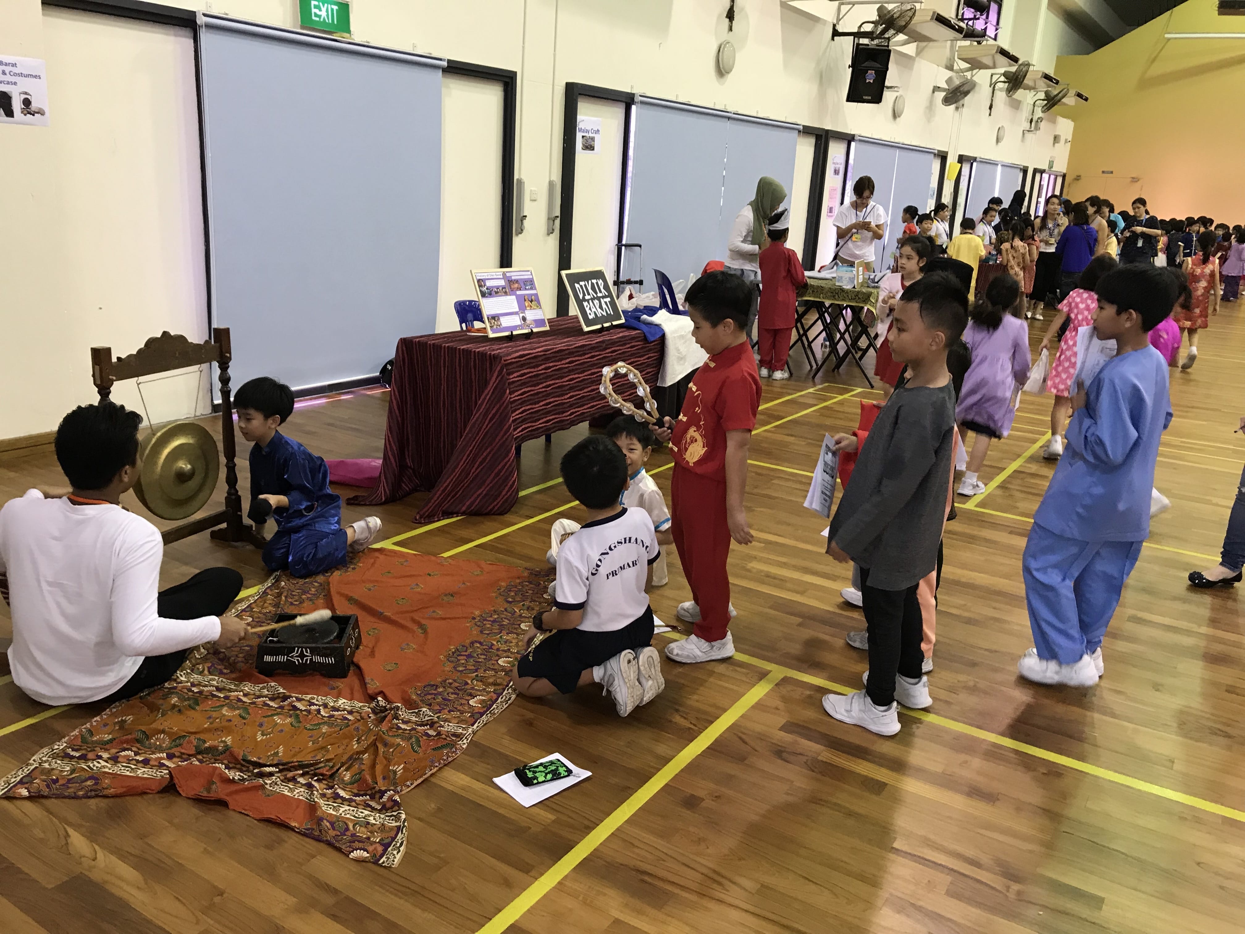 Children in traditional costumes explore a cultural fair in a school hall, with a gong and Dikir Barat display booth visible.