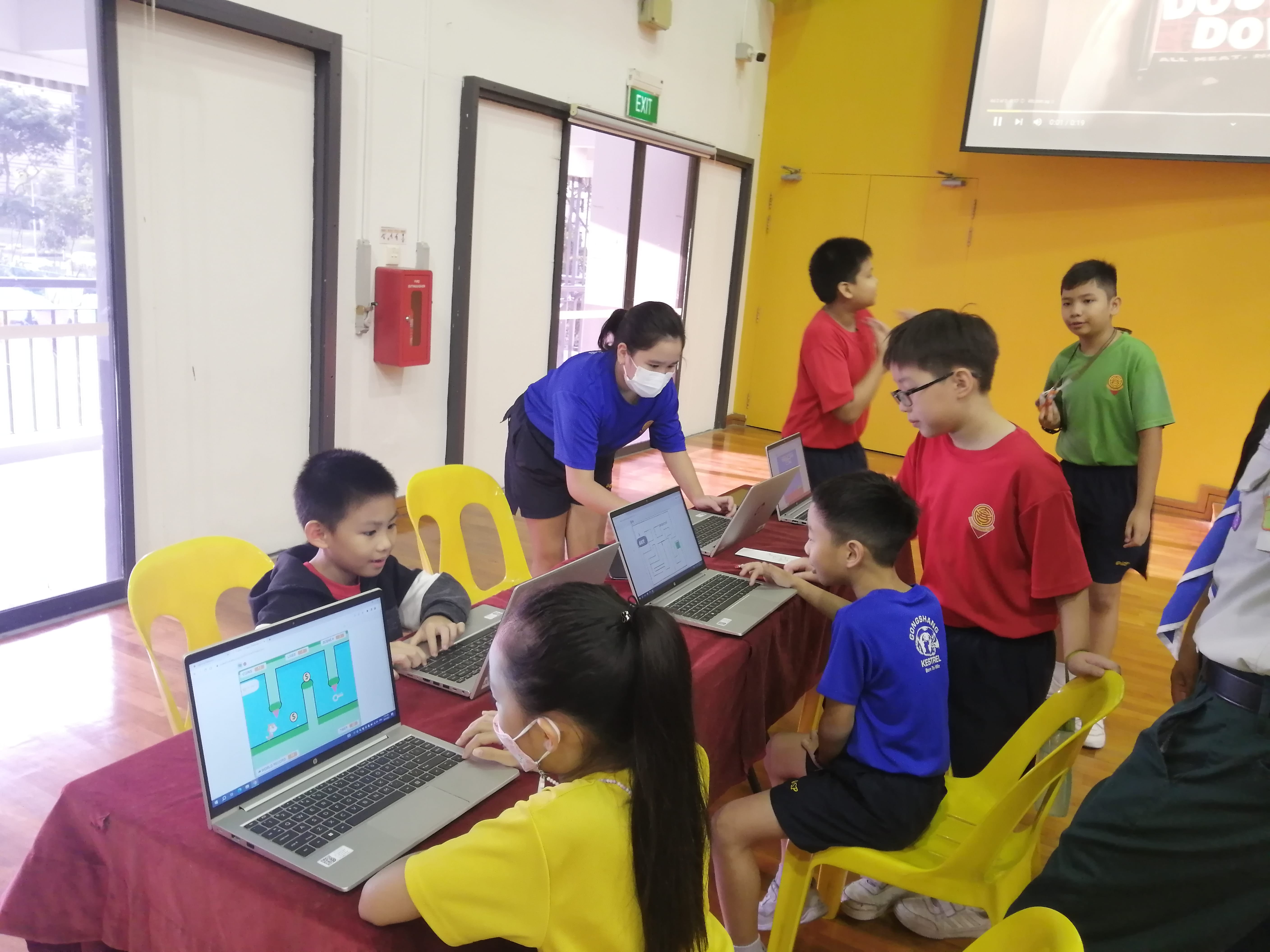 Children sit at a red table using laptops while a masked adult in blue leans over to assist them in a hall.